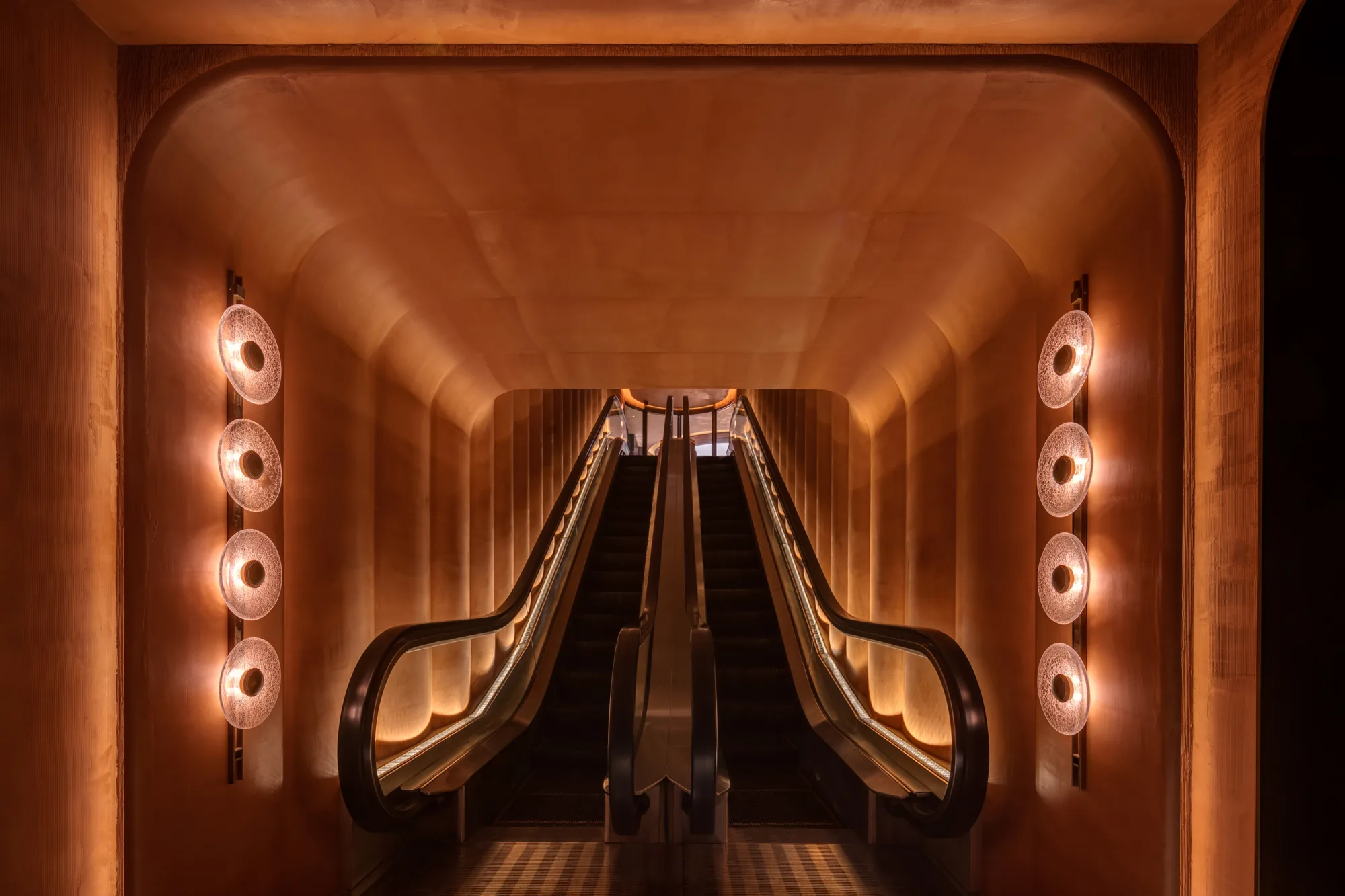 Copper-toned tunnel with curved walls and ceiling framing two side-by-side escalators. Circular wall lights line both sides, and warm strip lighting traces the edges.