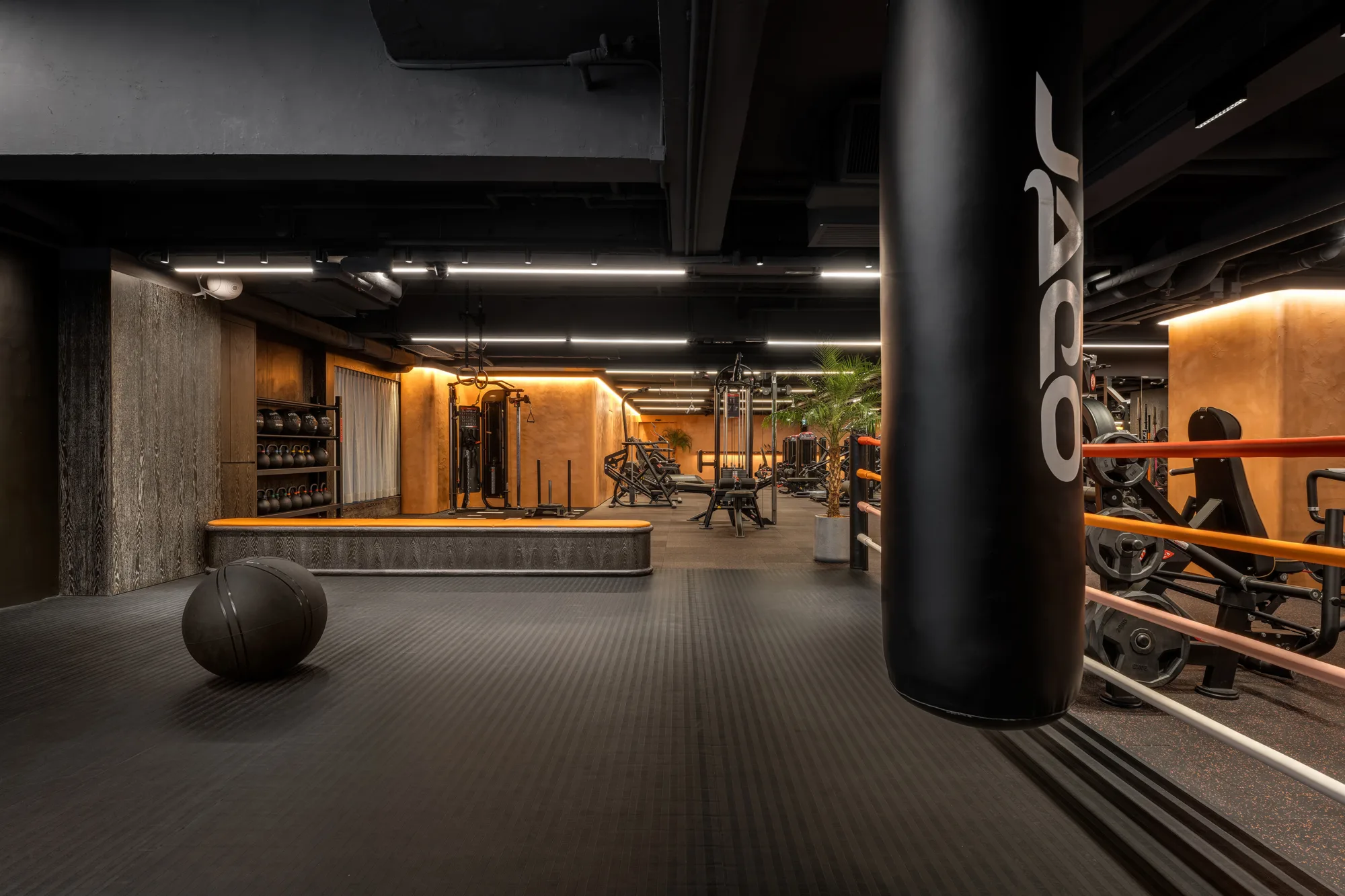 Modern gym interior with dark rubber flooring, a black punching bag with partial white lettering, strength machines, barbells, and kettle-bells on racks. Exposed ceiling pipes and strip lighting; orange-toned walls and a potted plant near equipment.