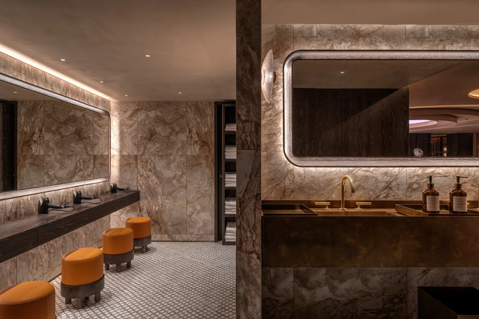 Marble-lined restroom with patterned floor tiles, large backlit mirrors, and a dark wood counter holding multiple hair dryers. Orange round stools line the vanity, and a metal sink with two soap bottles sits beside open shelving with folded towels.