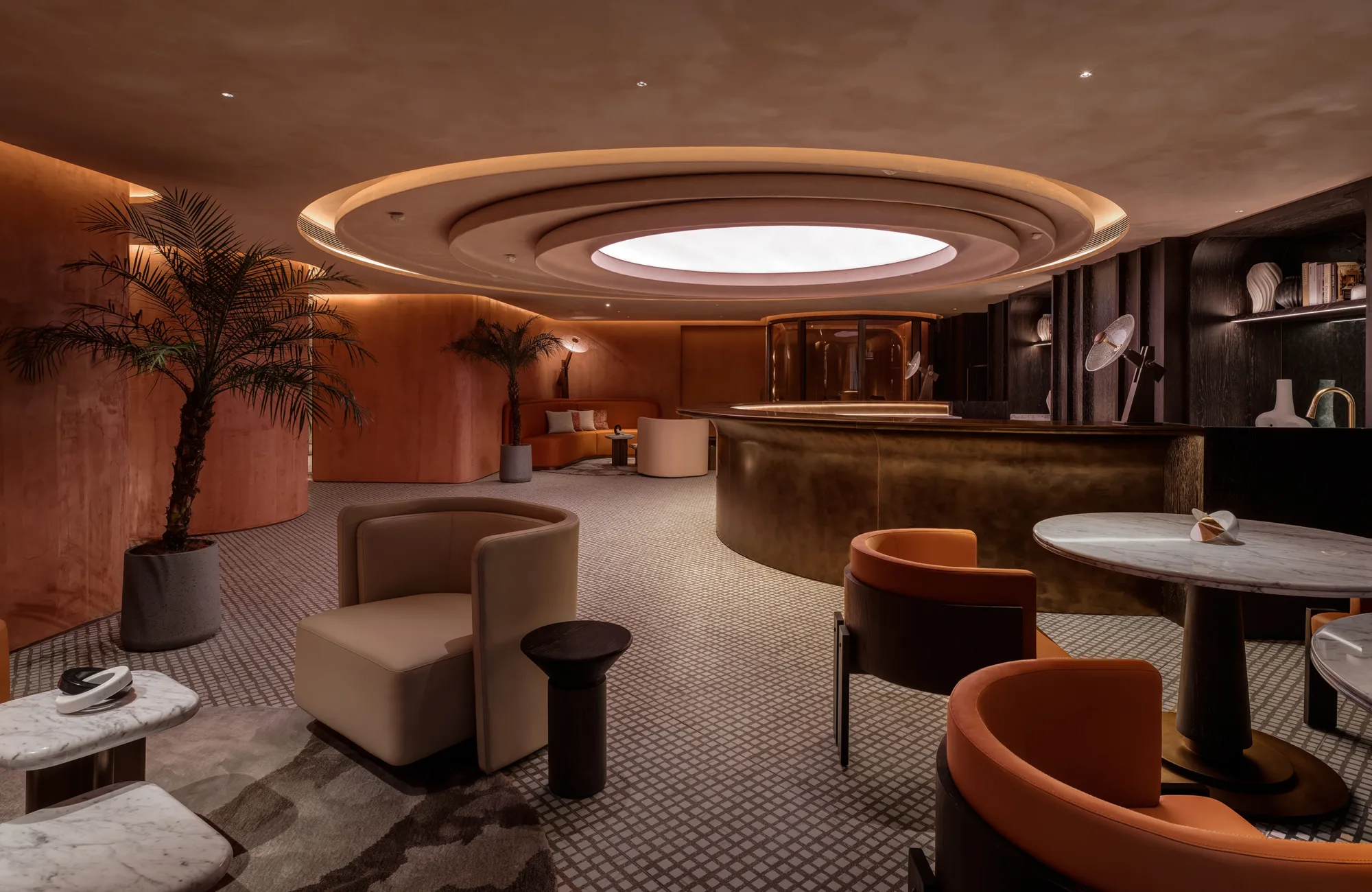 Modern lounge interior with curved copper-toned walls and ceiling, circular recessed light feature, marble tables, upholstered chairs, potted palm plants, and a rounded reception desk with shelves holding books and decorative objects.