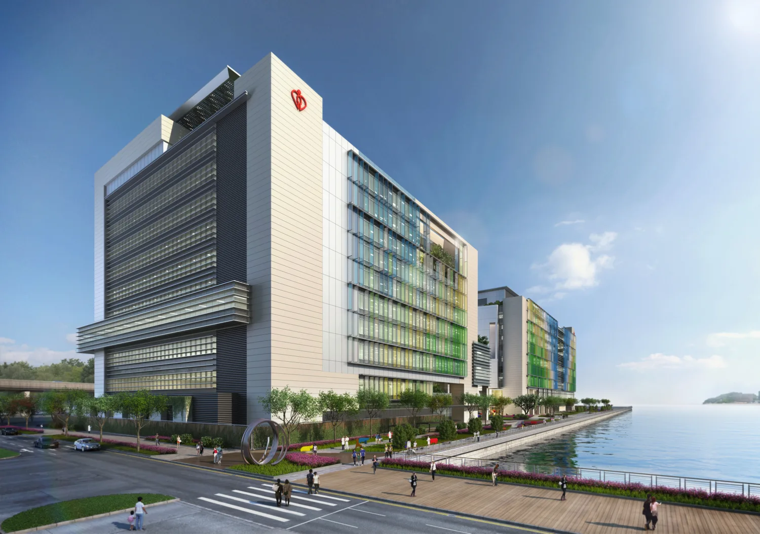 Large light coloured hospital building sitting on a waterfront. In the foreground is a road, zebra crossing and a line of trees separating this from the building