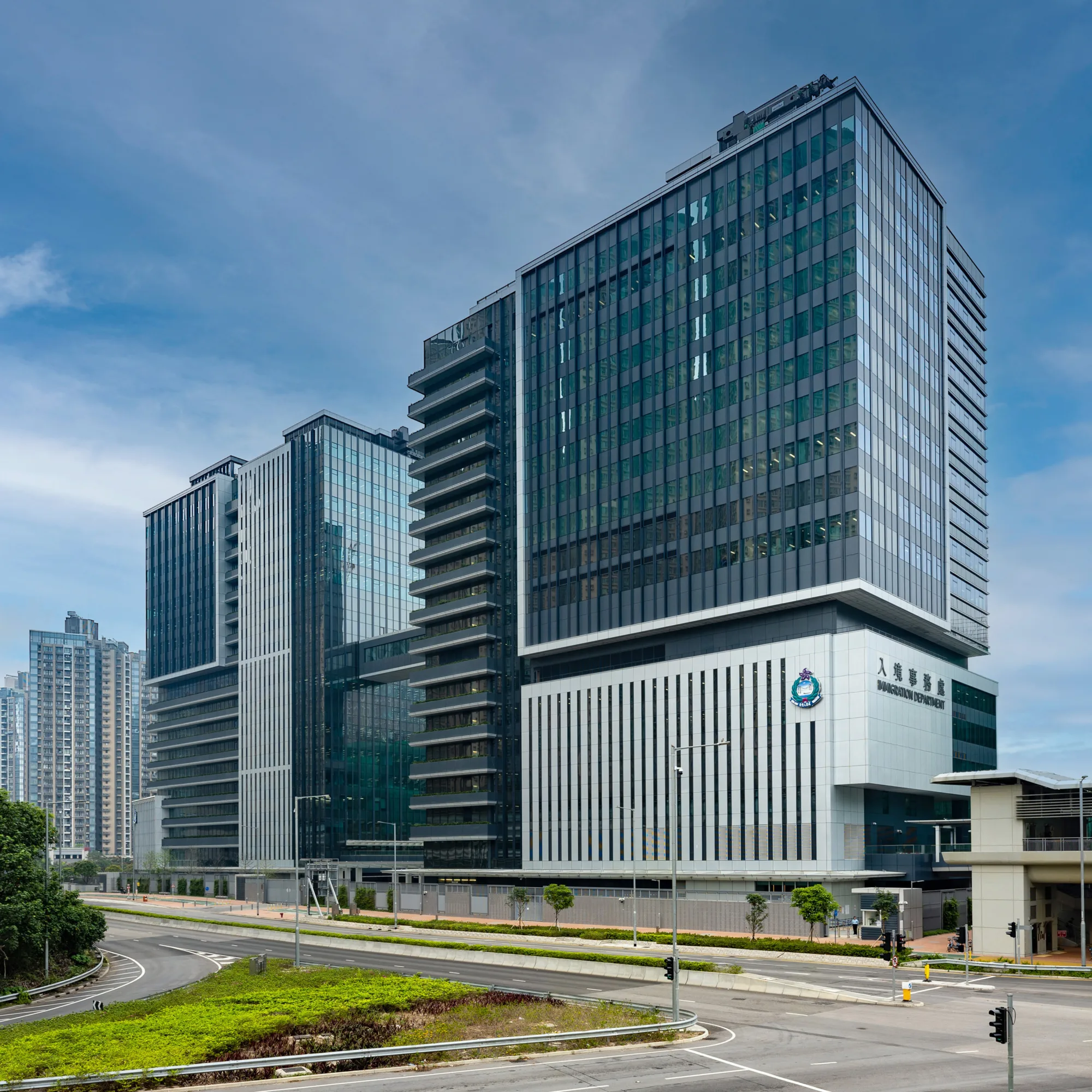 Multi-story office building with reflective glass windows and vertical white stripes on lower floors, located beside a wide empty road with green landscaping and a clear blue sky.