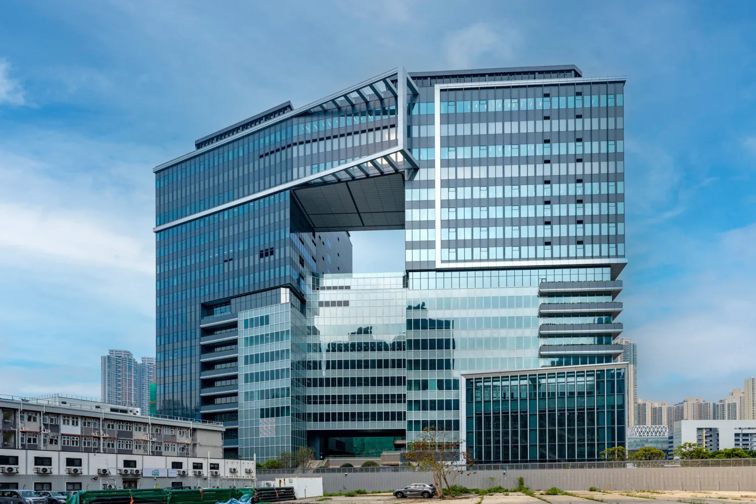 Multi-story glass office building with an open central section, reflective windows, blue sky background, construction site in foreground, and nearby low-rise buildings with greenery.
