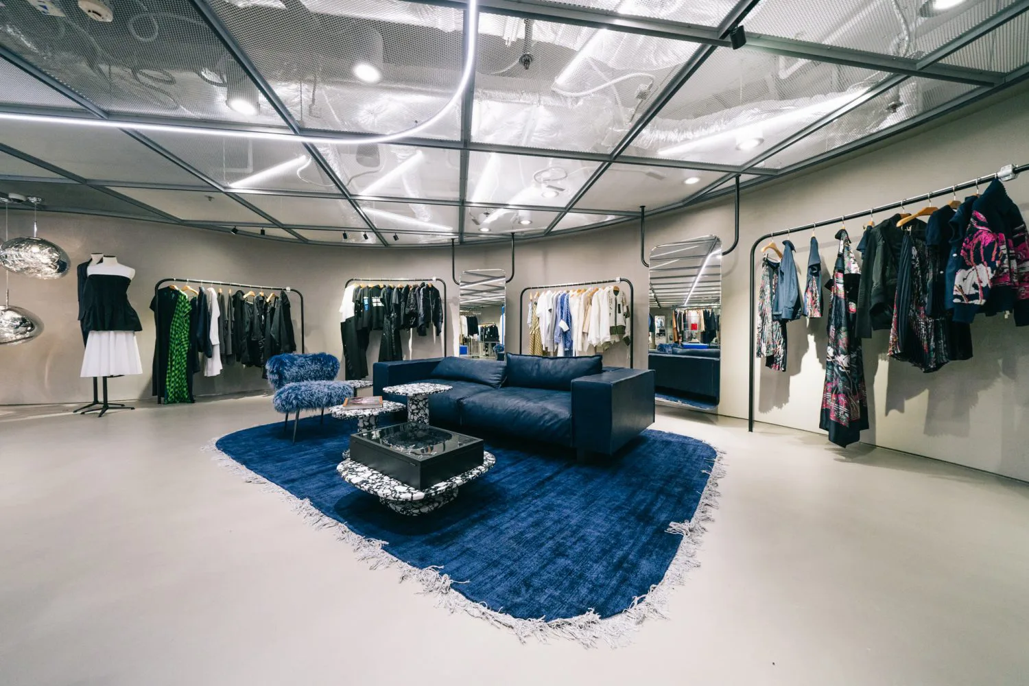 The clothing display area, a sofa was placed in the centre and clothes are displayed on the wall behind the sofa. Tailor-made downlight fixture can be seen on the metal mesh ceiling.