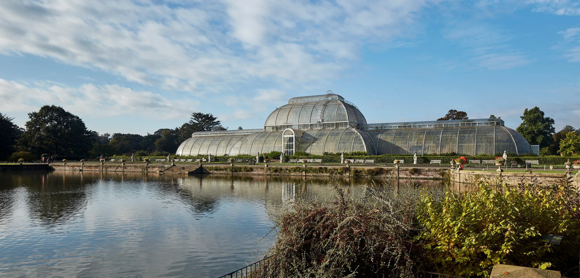 The glass house conservatory with a lake in the foreground and a blue sky with light cloud coverage