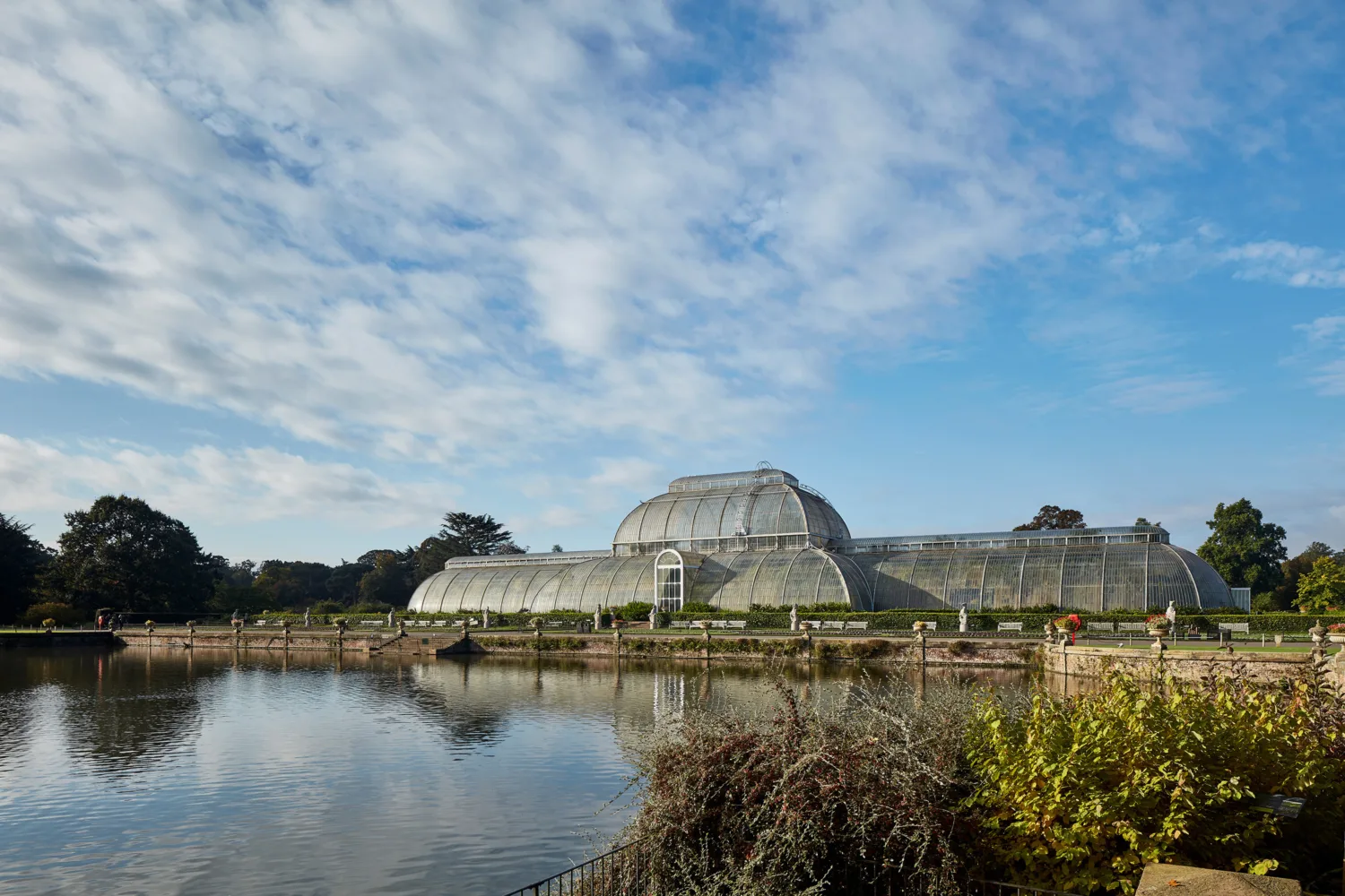 The glass house conservatory with a lake in the foreground and a blue sky with light cloud coverage