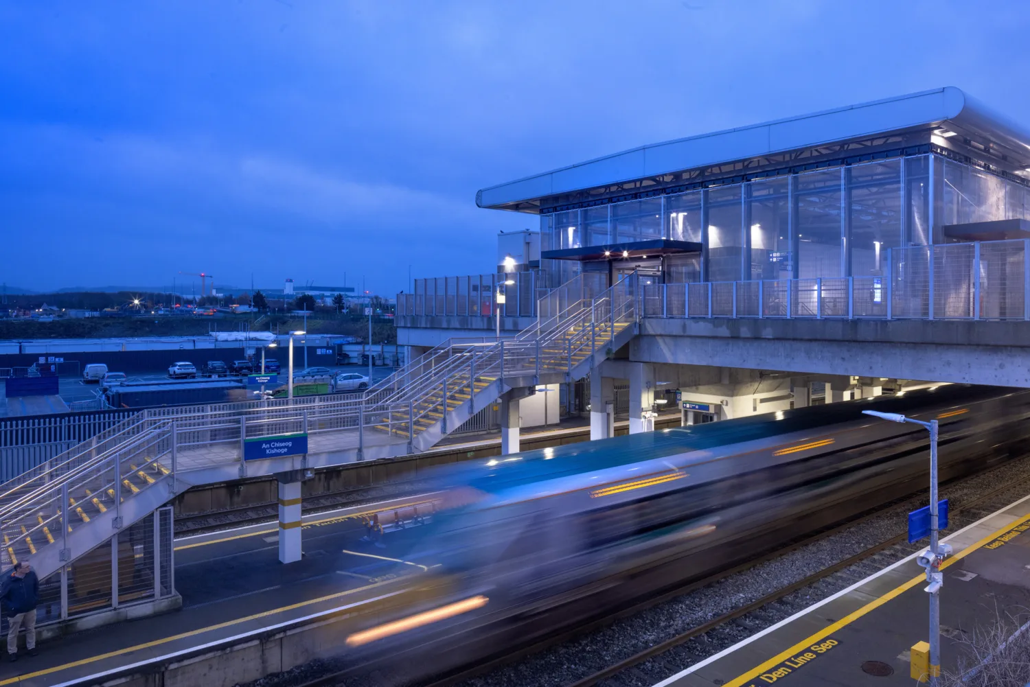 Moving train passes through illuminated Kishoge station at dusk.
