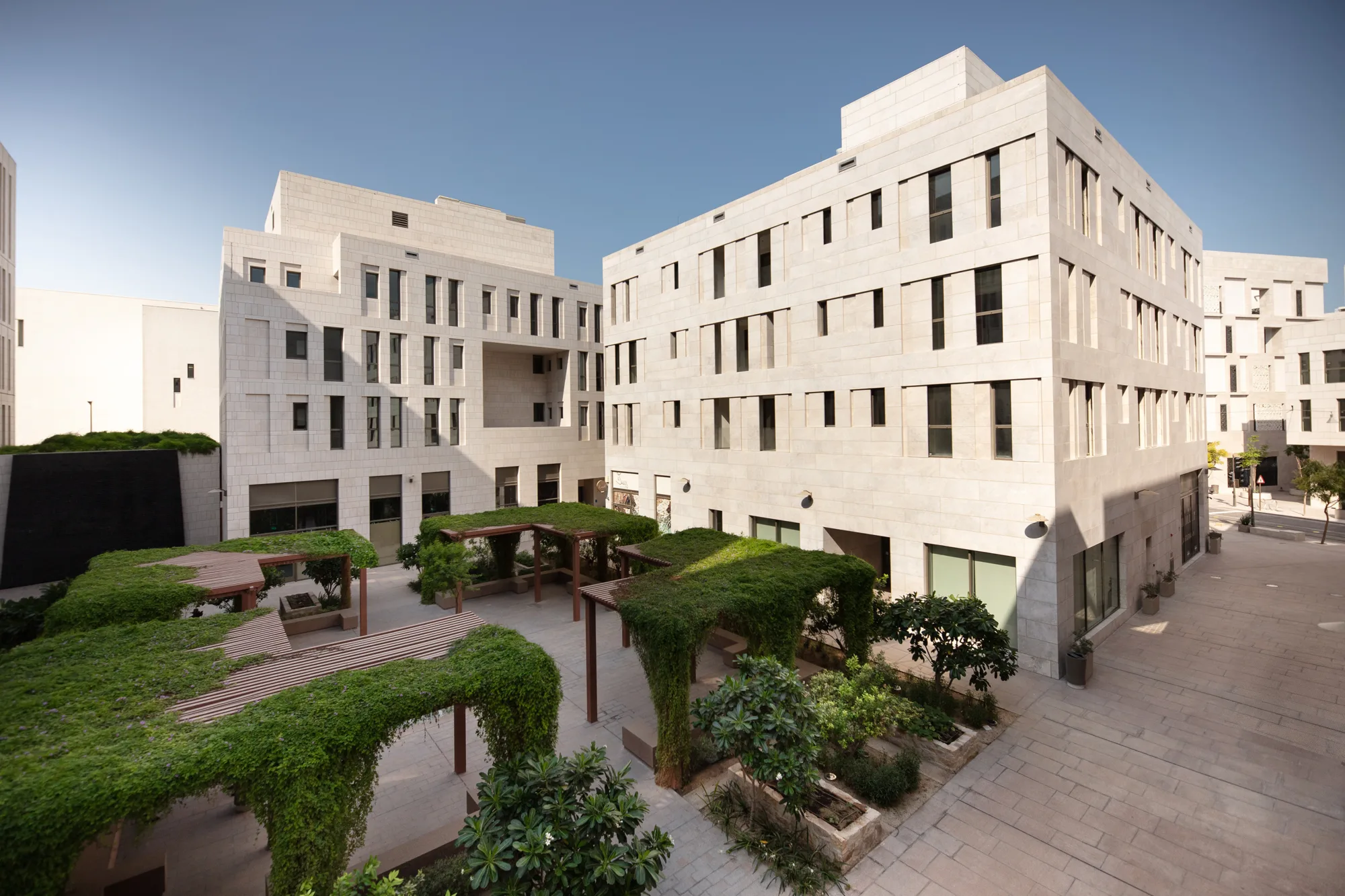 Beige stone buildings frame a landscaped courtyard with pergola structures covered in greenery, paved walkways, raised planters, small trees, and shaded seating niches under clear daylight sky.