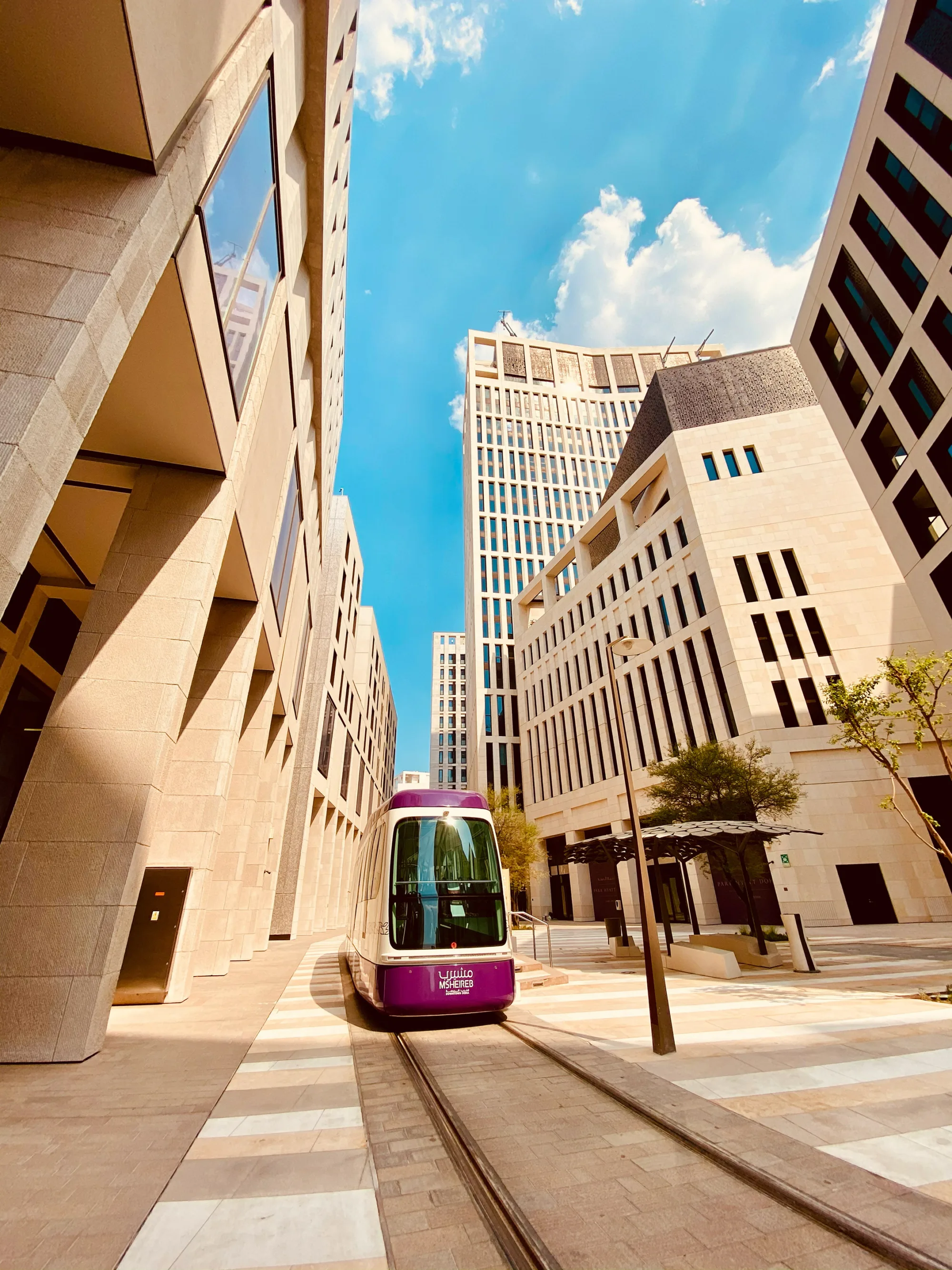 Street-level view of a modern urban district with a purple-front tram on tracks between beige stone buildings. Tall facades frame a blue sky with clouds; shaded walkways, trees, and patterned paving line the corridor.