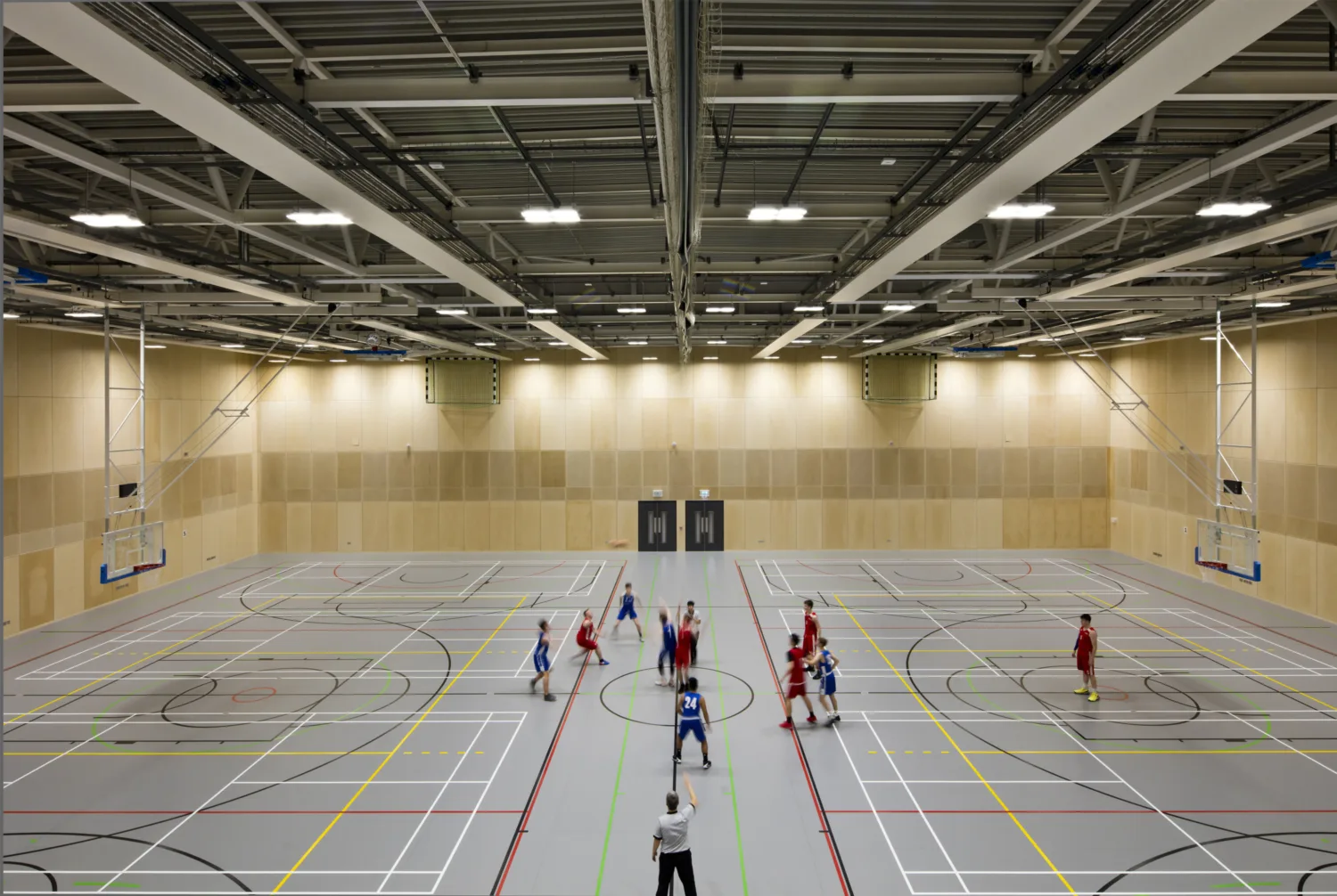 Modern sports hall interior with light wooden walls and a floor with various coloured lines painted, a group of people are laying a sports game in the centre of the hall