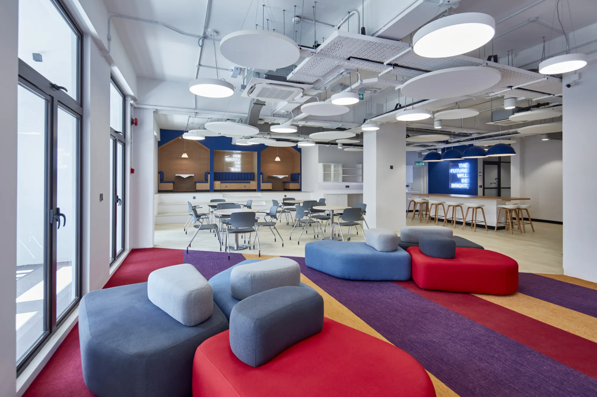 Brightly lit space with multi-coloured sofas on carpeted floor; background features work space with round tables, work booths, and rectangular table