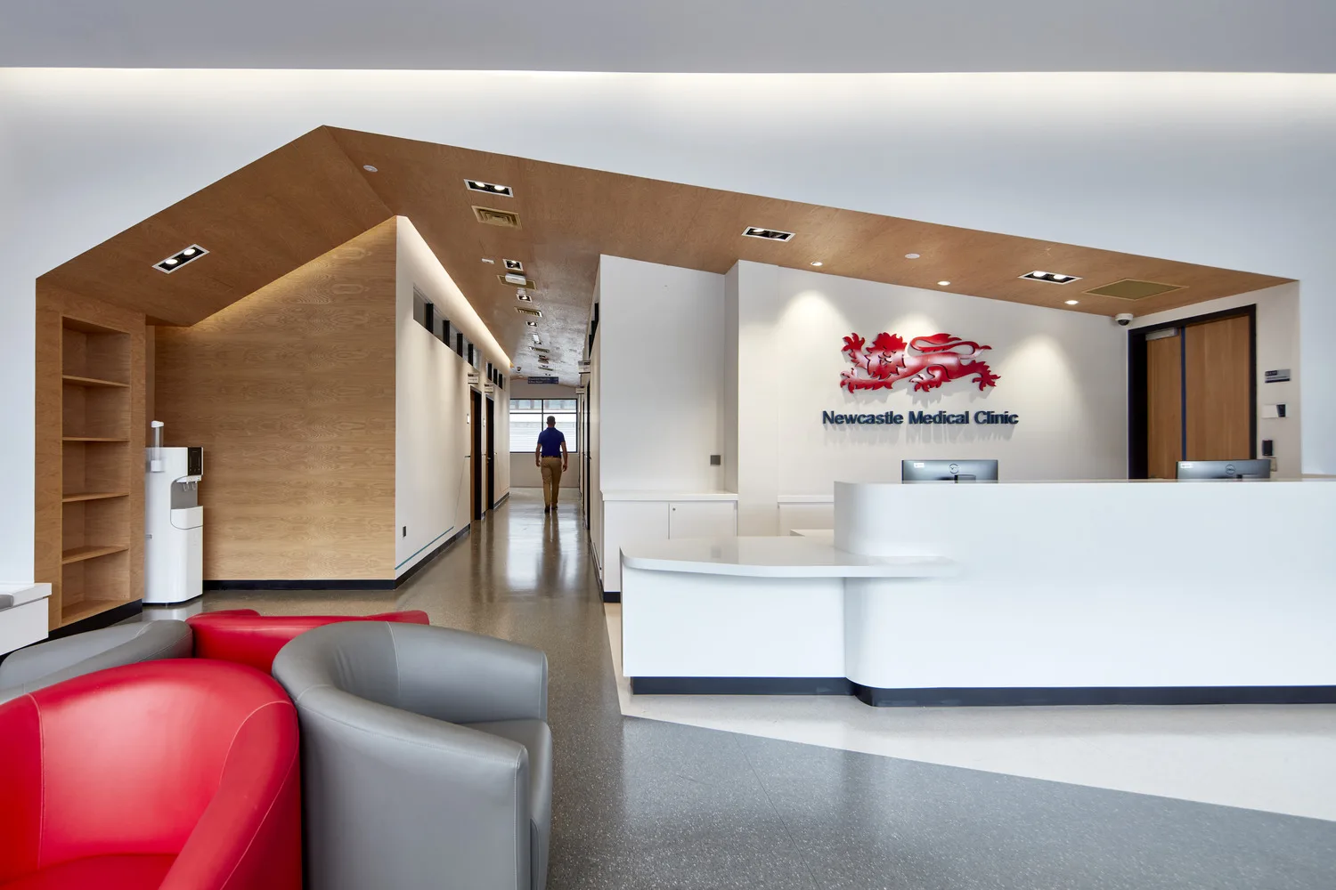 Open area featuring red / grey armchairs and reception counter with NUMed logo beside hallway underneath vaulted ceiling