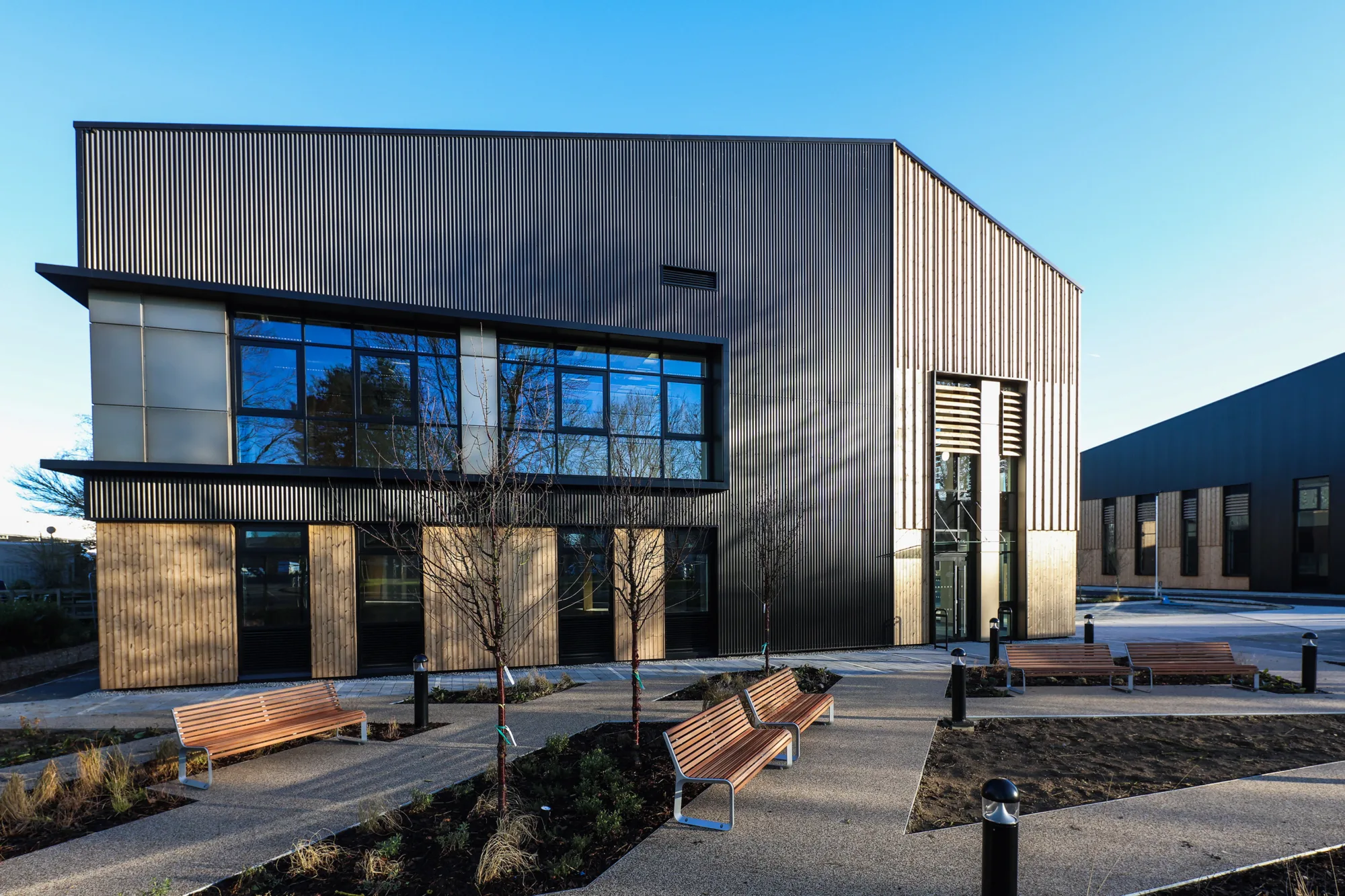 Contemporary commercial building with timber cladding, dark metal façade, large glazed windows and landscaped external seating areas.