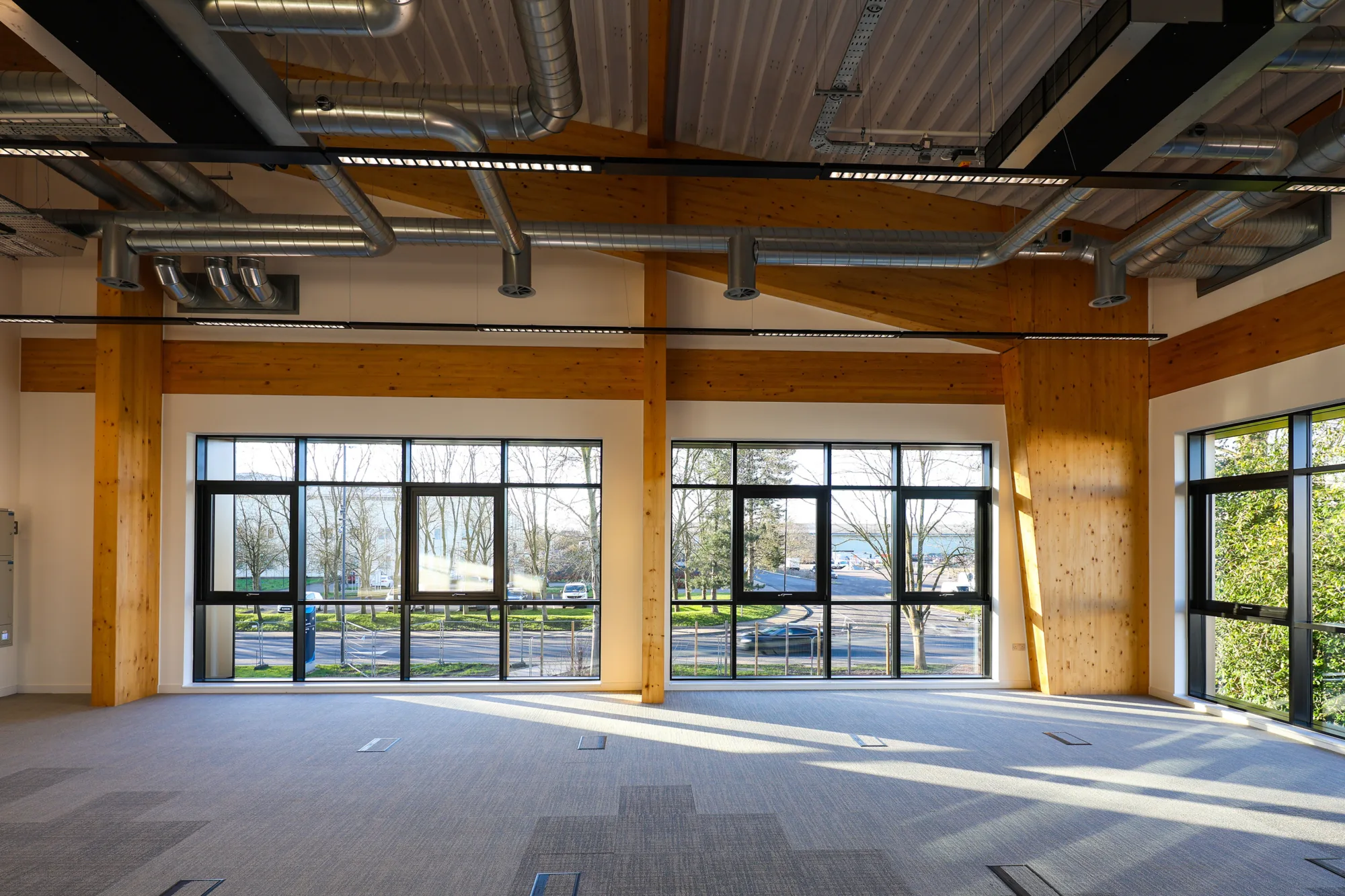 Large open-plan interior with exposed timber structure, visible ventilation ductwork, carpeted flooring and floor-to-ceiling glazing admitting natural daylight.