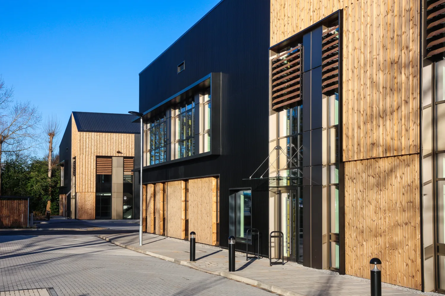 Commercial building façade combining timber cladding, dark metal panels and extensive glazing along a paved pedestrian route.