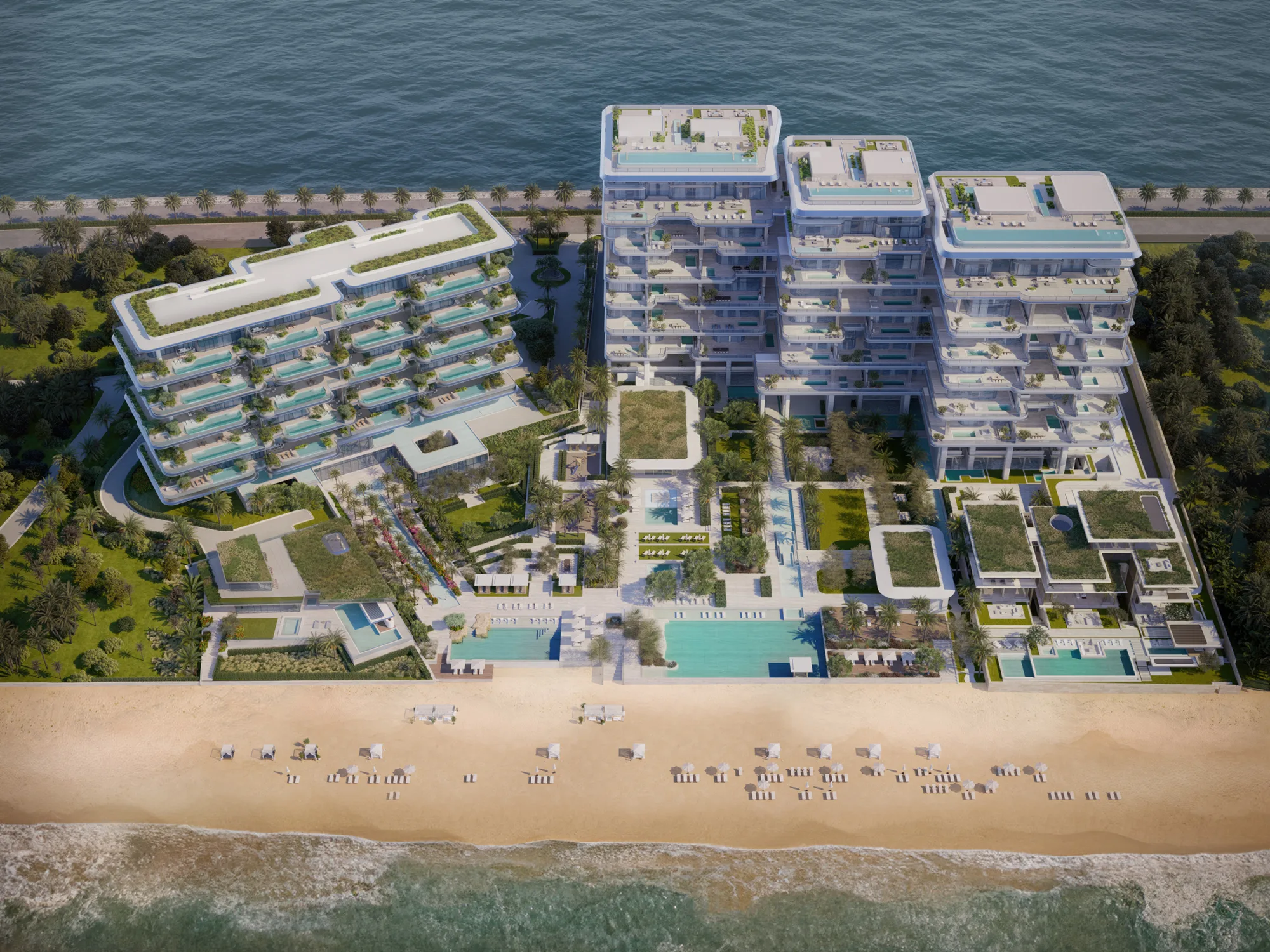 Aerial view of a beachfront complex with several modern terraced buildings featuring layered balconies, rooftop greenery, and pools. Landscaped gardens lead to a sandy beach with arranged loungers beside calm sea water.
