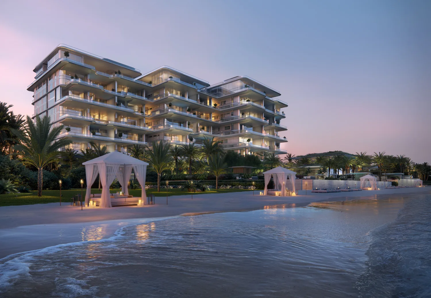Beachfront view of a multi-level modern building with layered balconies and warm interior lighting at dusk. Palm trees, cabanas, and a sandy shore frame calm water in the foreground.