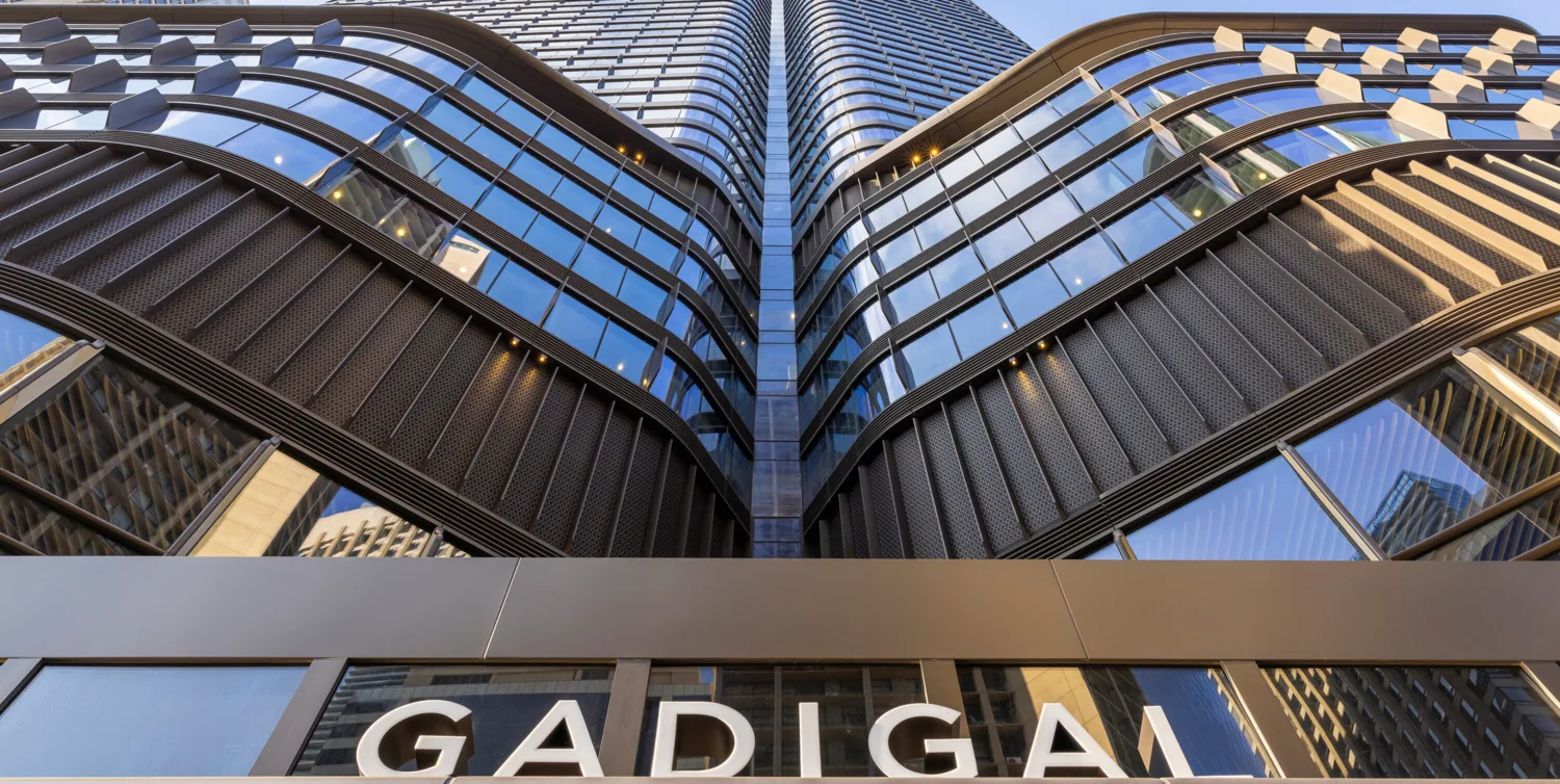 Low-angle view of a modern high-rise building with curved, layered glass and metal façade reflecting the sky and nearby structures. The word “GADIGAL” is visible on a sign at the bottom of the image.