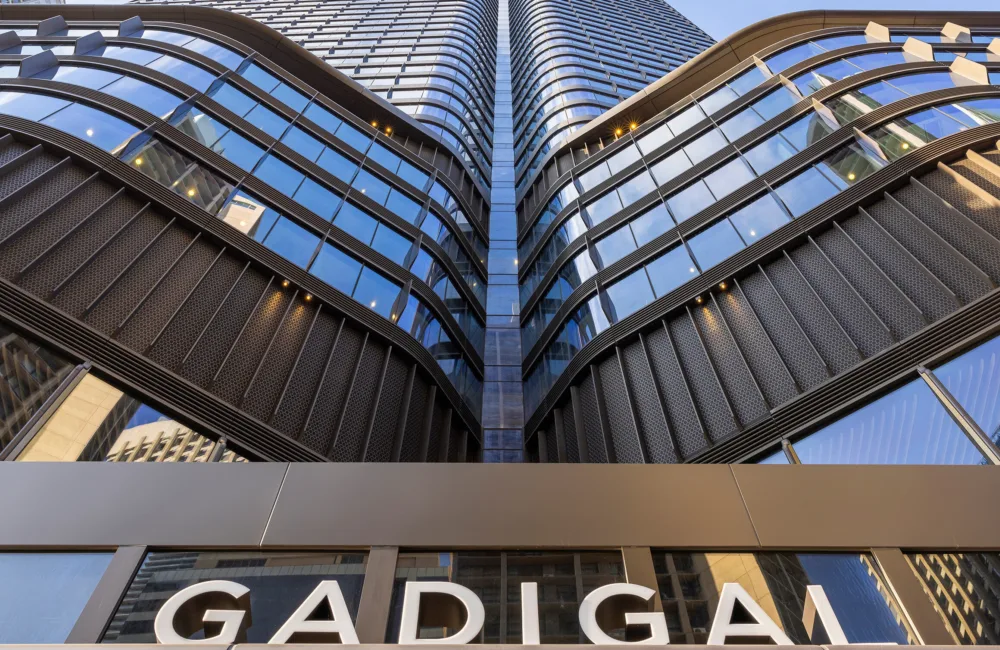 Low-angle view of a modern high-rise building with curved, layered glass and metal façade reflecting the sky and nearby structures. The word “GADIGAL” is visible on a sign at the bottom of the image.