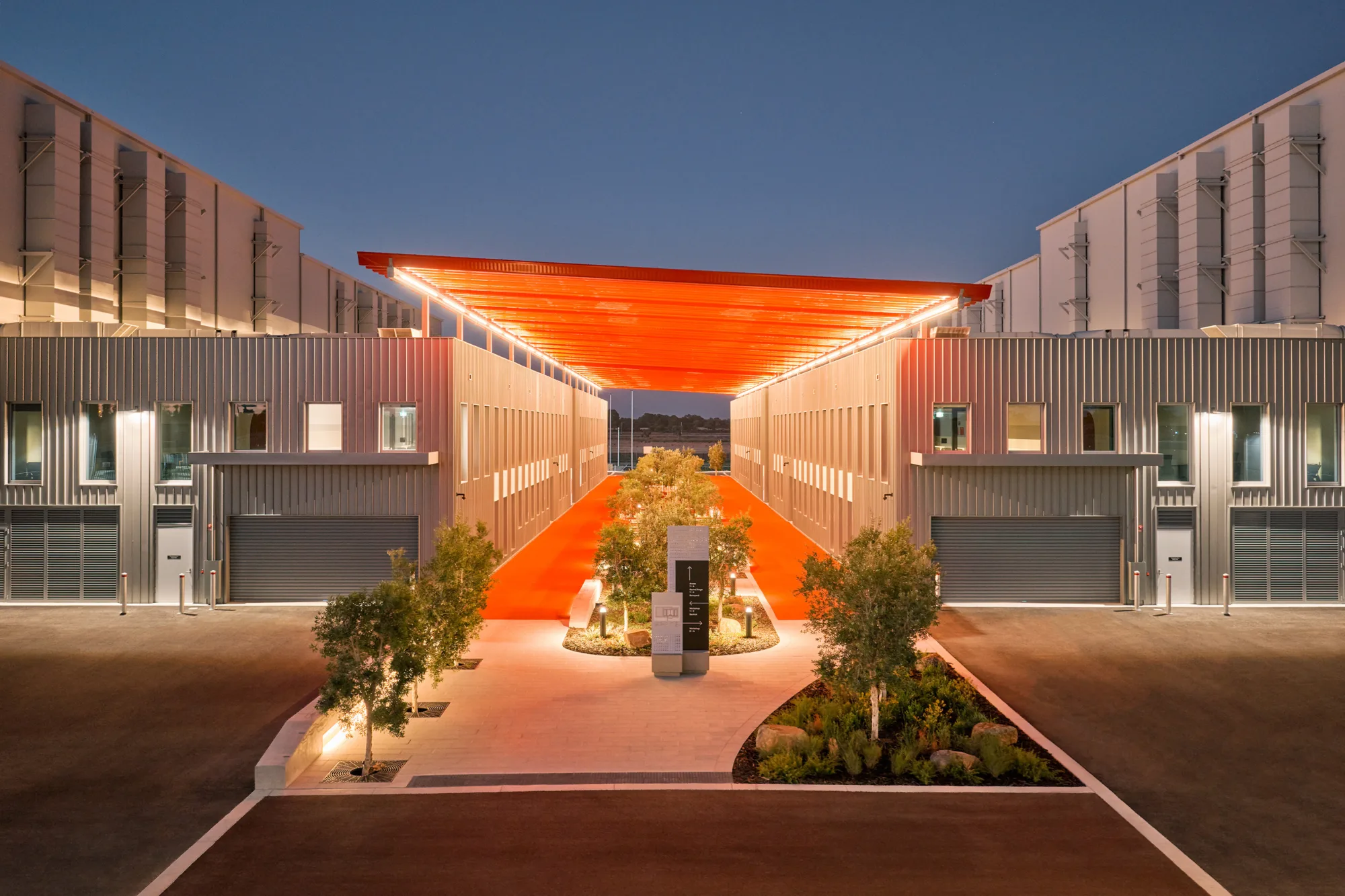 Symmetrical view between two industrial buildings with metal facades and rows of windows. A bright orange illuminated canopy spans overhead above a central landscaped walkway with trees, lighting, and a signboard; paved roads border each side.