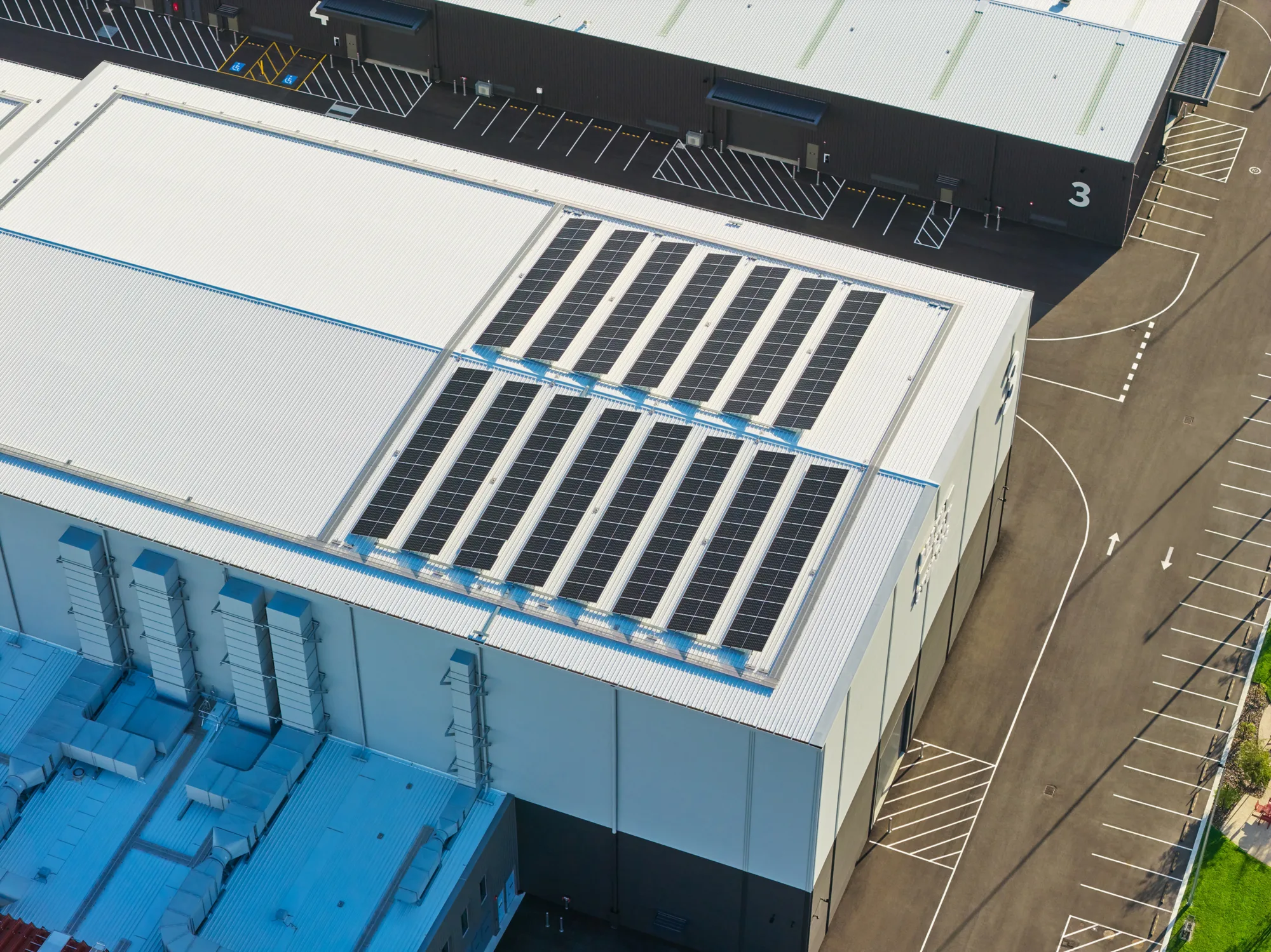 Aerial view of industrial buildings with white roofs fitted with rows of solar panels. Marked parking bays, road lines, and large wall numbers “3” and “4” are visible around the paved site.