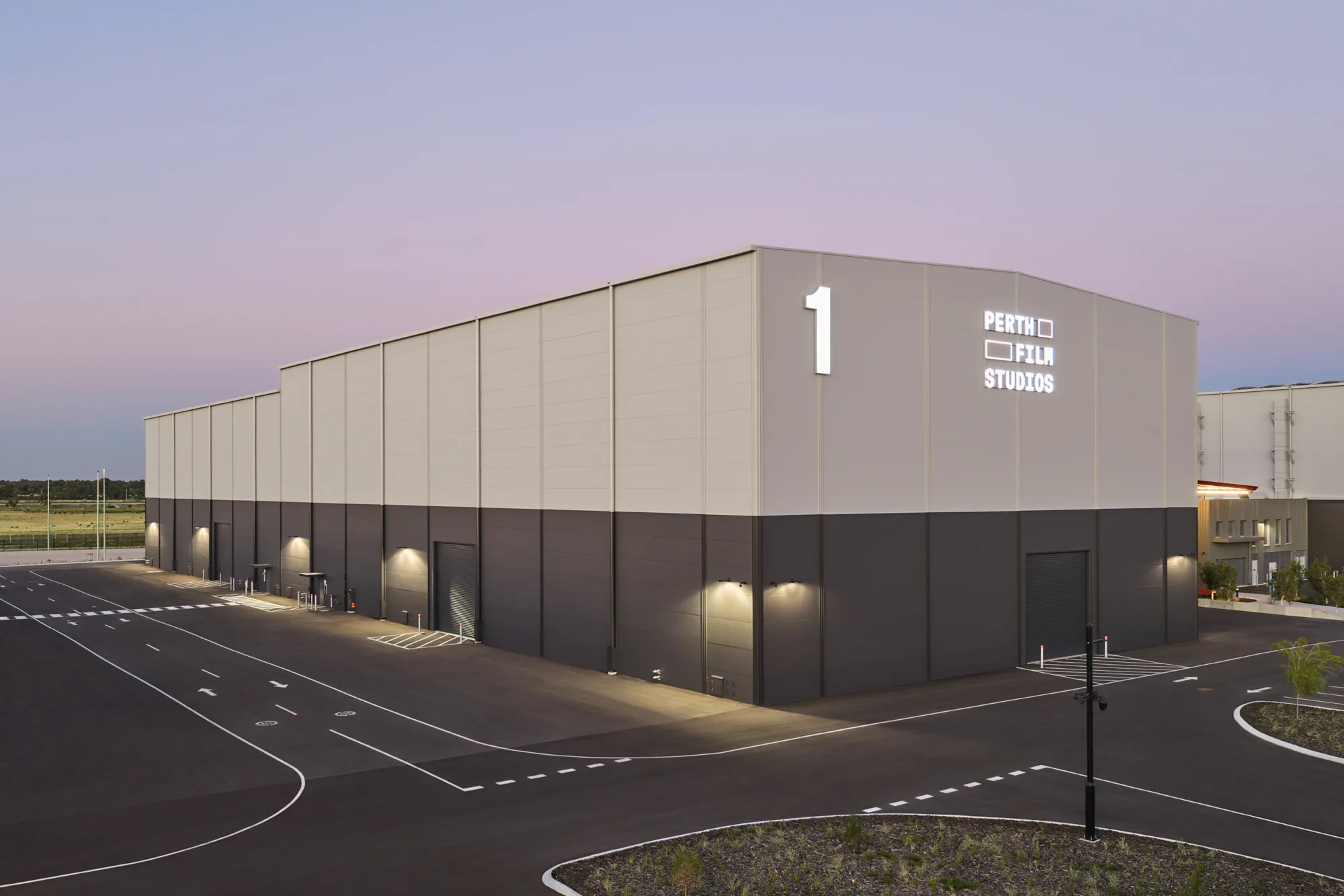 Large rectangular industrial building marked “1” with illuminated text “PERTH FILM STUDIOS” on the upper wall. Paved roads, lane markings, roller doors, and exterior lights surround the structure under a dusk sky.