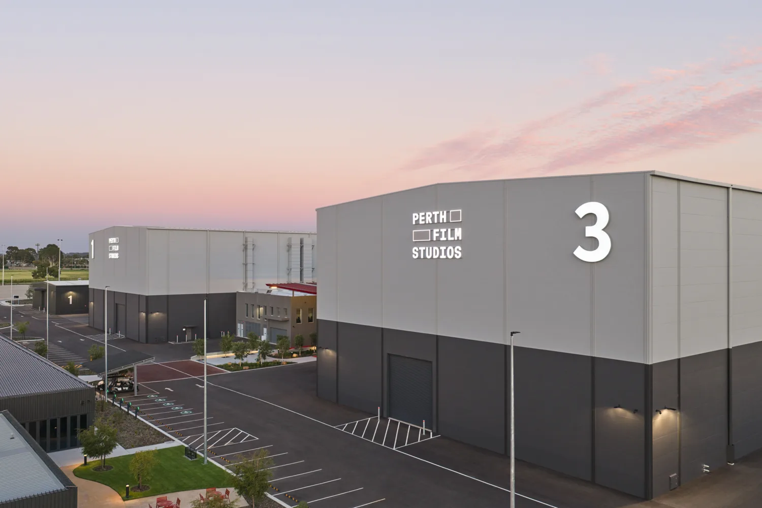 Large industrial buildings at a studio complex, with illuminated text “PERTH FILM STUDIOS” and large numbers “3” and “1” on exterior walls. Paved parking areas, marked bays, landscaped greenery, and streetlights sit under a pink dusk sky.