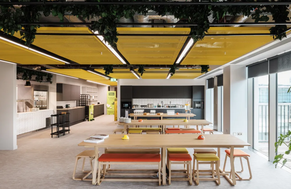 Staff eating and coffee area, yellow acoustic panelling ceiling with wooden tables with orange and yellow seating, as well as black and white units for the kitchen area