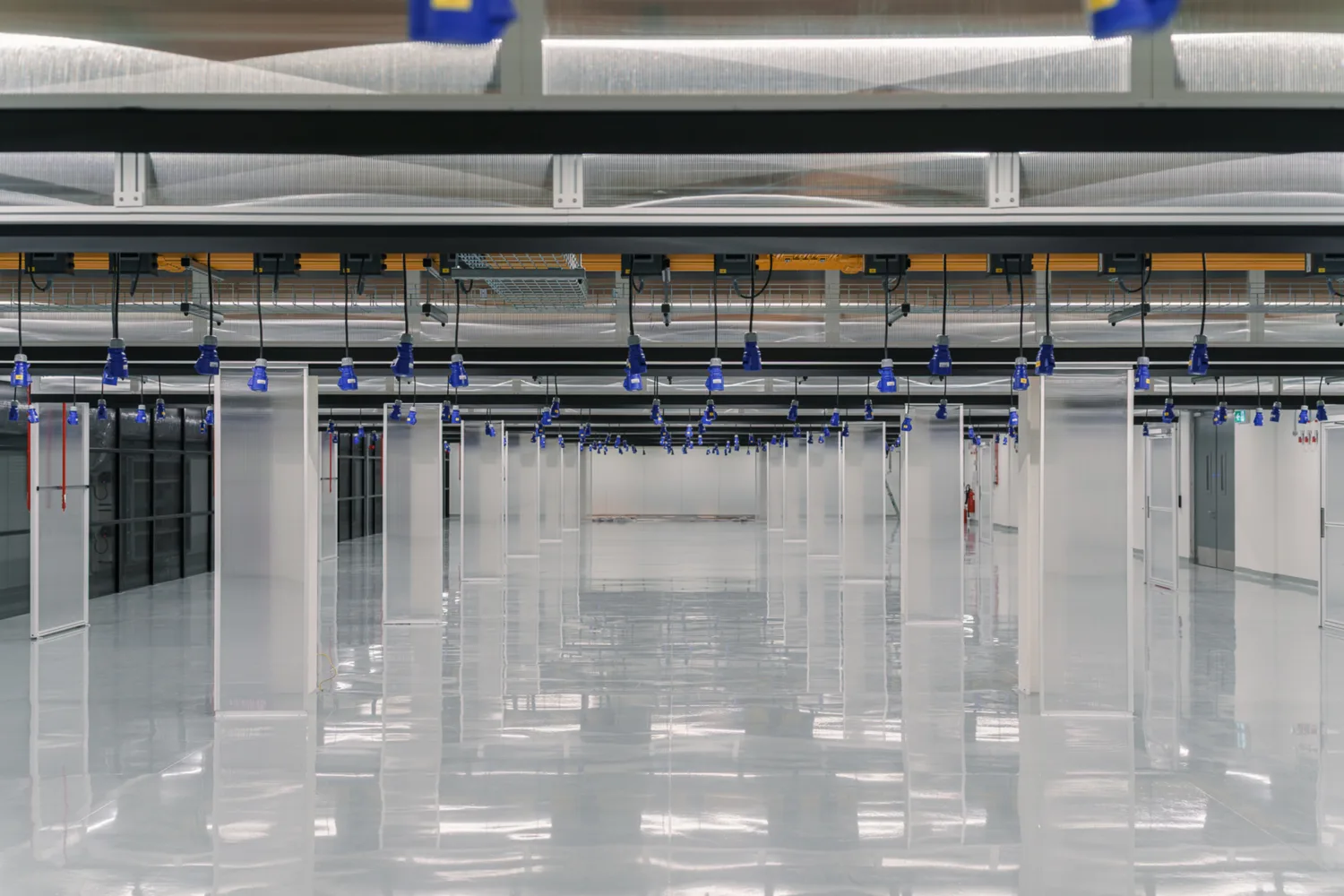 Large empty industrial room with a reflective floor, white columns evenly spaced, and a ceiling featuring blue hanging light fixtures and exposed ventilation ducts.