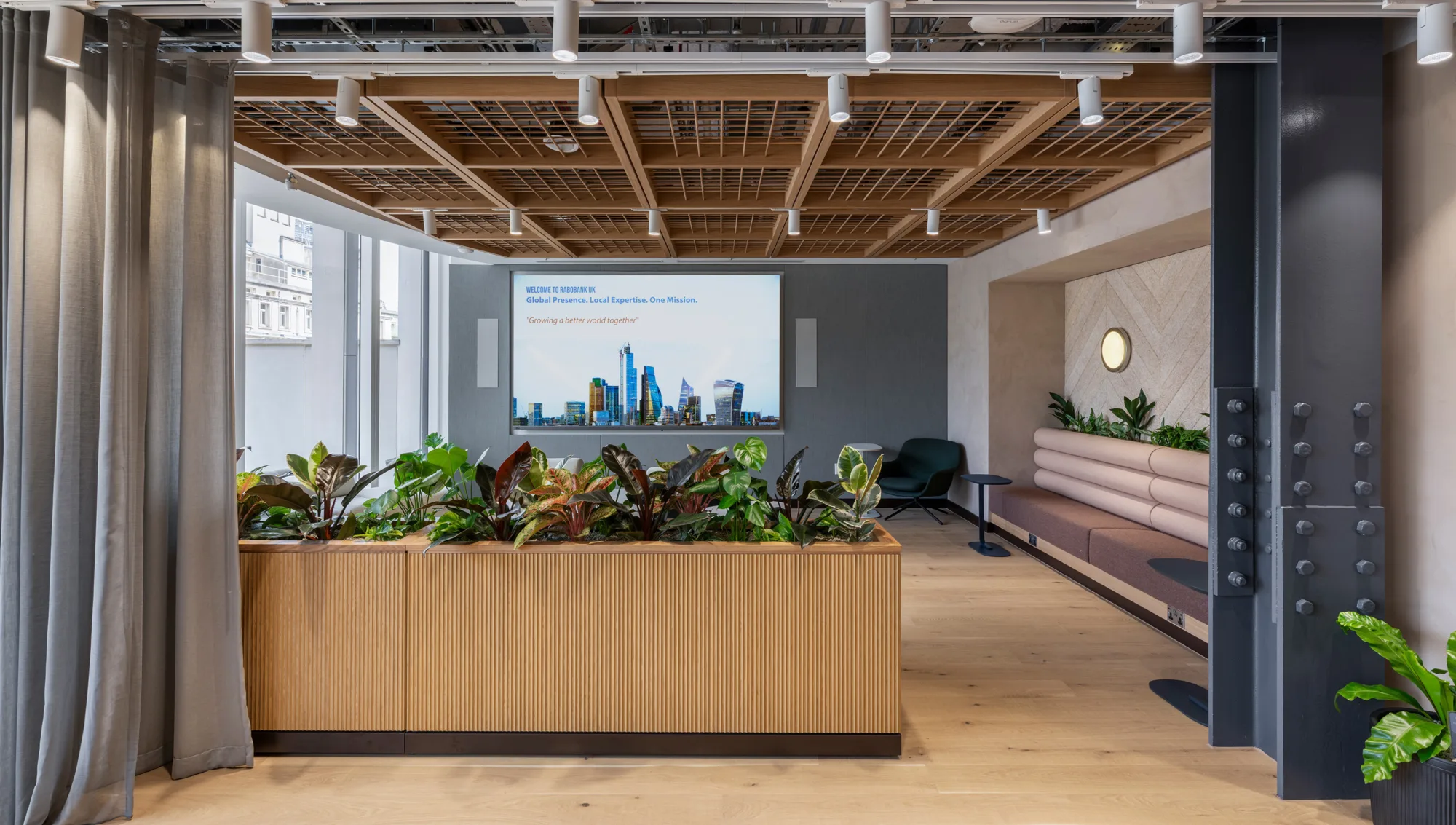 A modern interior space with a large planter of mixed green and red plants, wood flooring, a cushioned bench along the right wall, and a screen displaying city skyline graphics and text at the back.