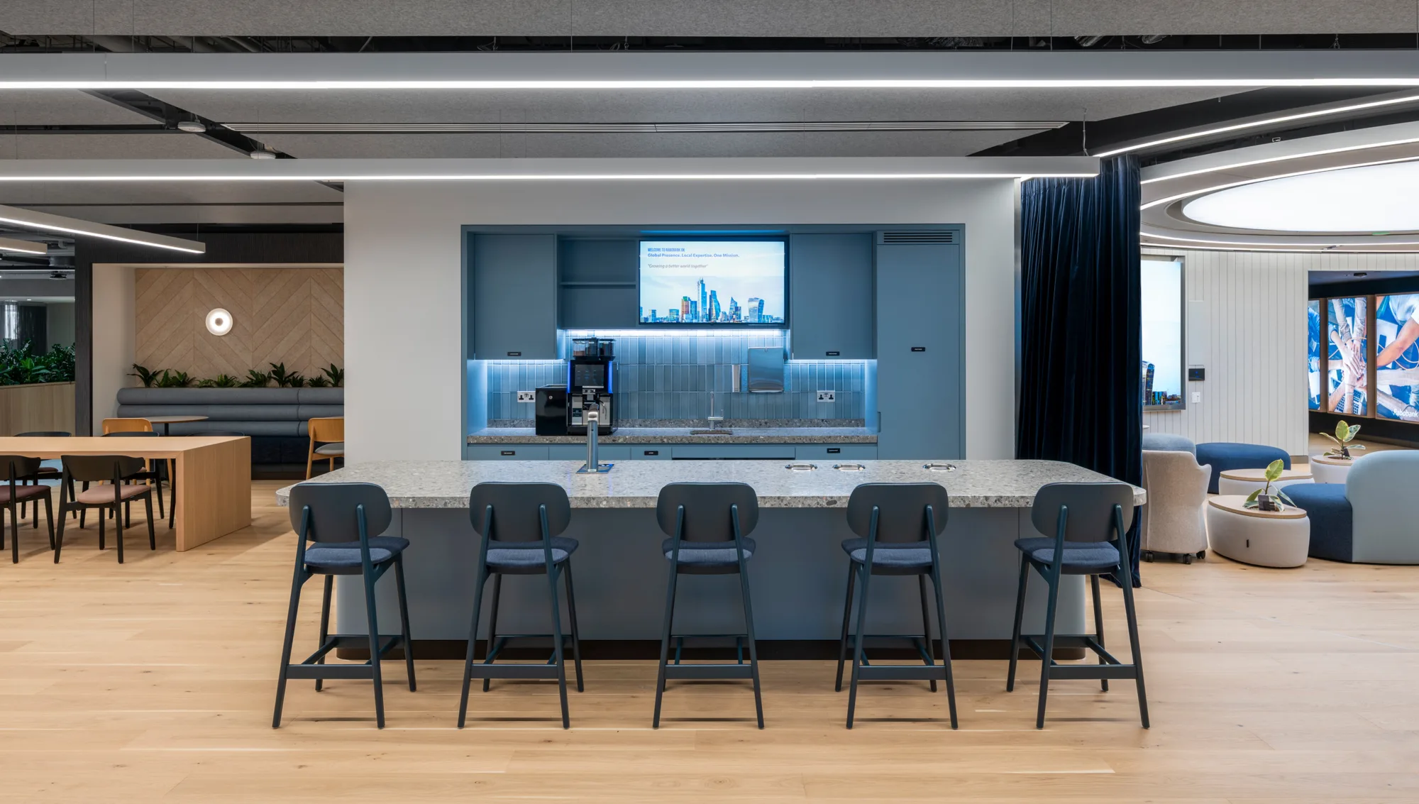 A modern kitchen area with a long counter, five bar stools, and built‑in appliances. A wall‑mounted screen displays a cityscape graphic above the counter. Surrounding areas include tables, seating, plants, and light wood flooring.