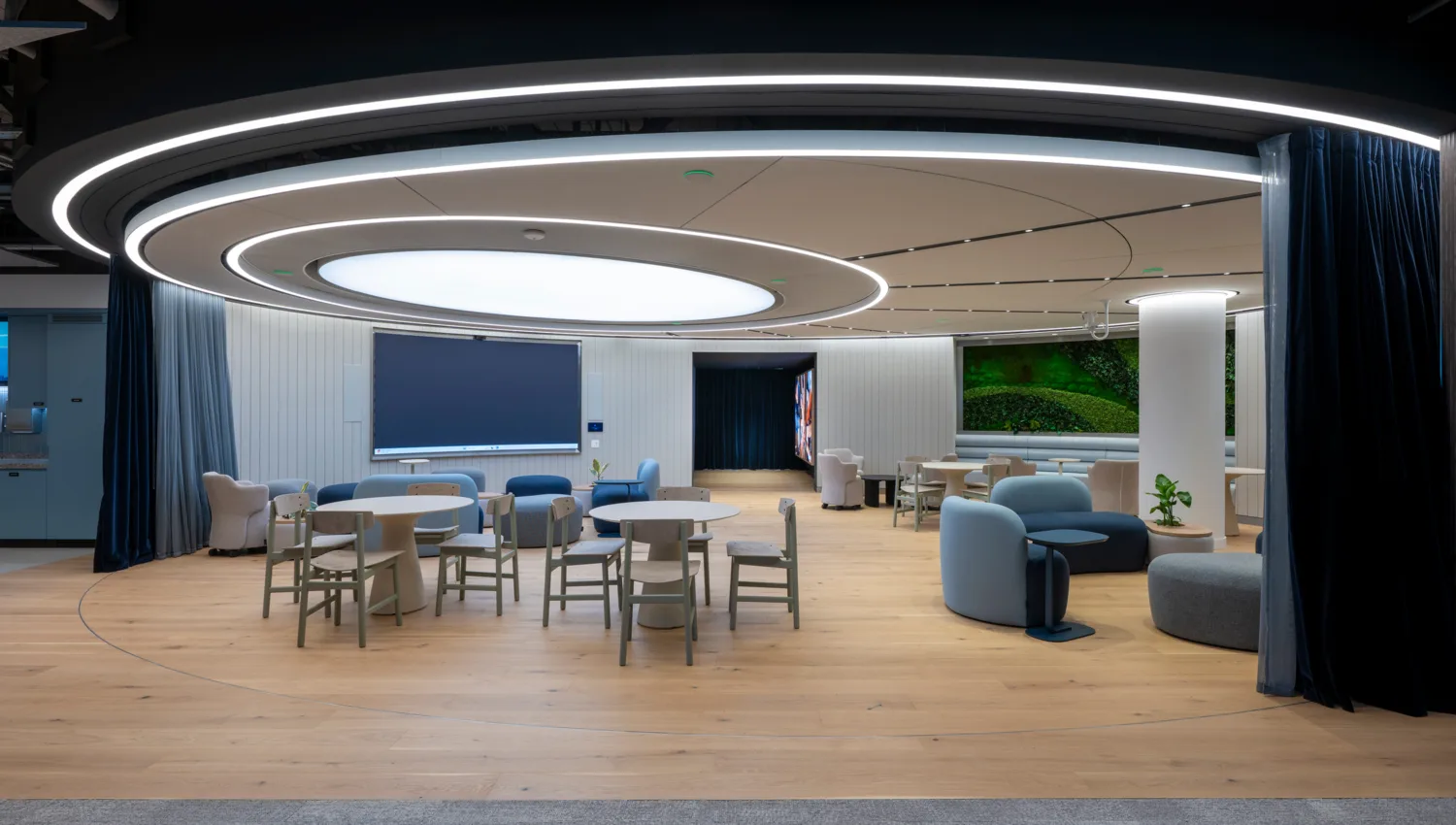 A modern open room with round ceiling lights, light wood flooring, and several tables with chairs. Lounge seating, a large blank screen, blue curtains, and a wall with greenery are visible throughout the space.