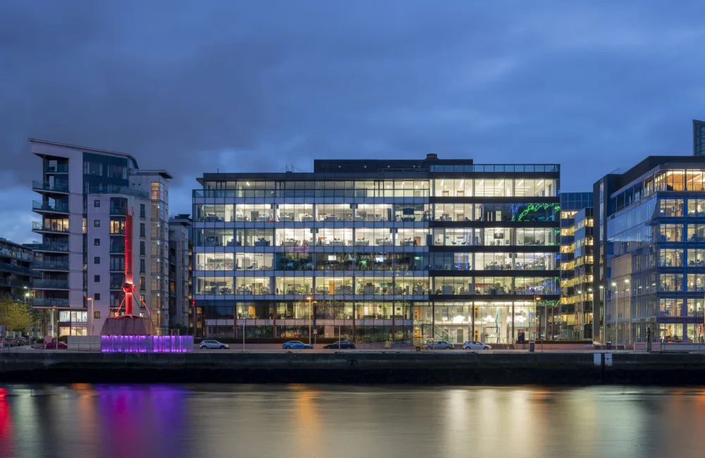 Cluster of buildings from the side in front of a river, brightly lit from the inside against a blue evening sky