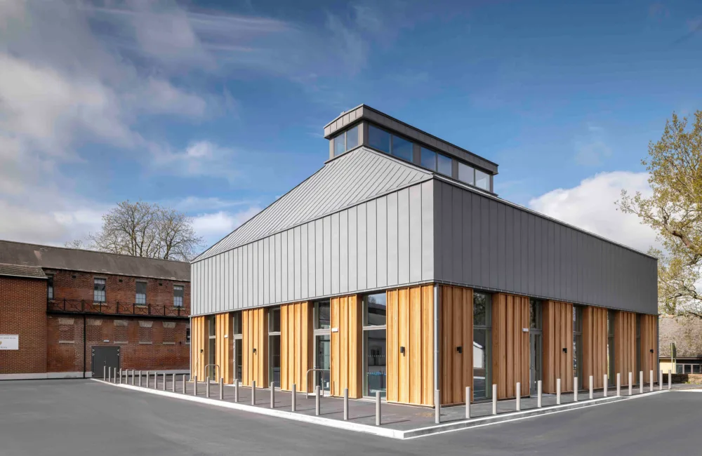 Exterior view of the Royal Military Academy Sandhurst Music Facility - a modern building with grey metallic panels, large glass windows, and wooden accents