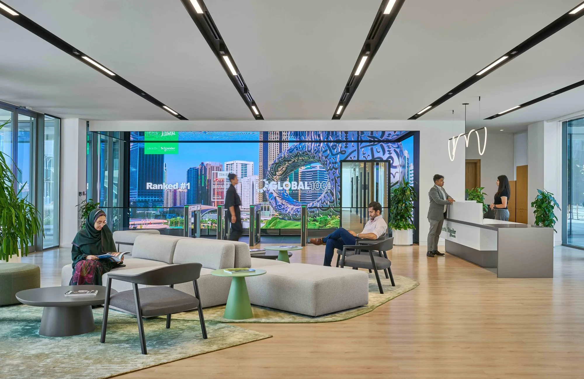 A modern lobby with lounge seating, low tables, and large digital screens showing city visuals. Several people sit, walk, or speak at a reception desk. Floor‑to‑ceiling windows and plants brighten the space.
