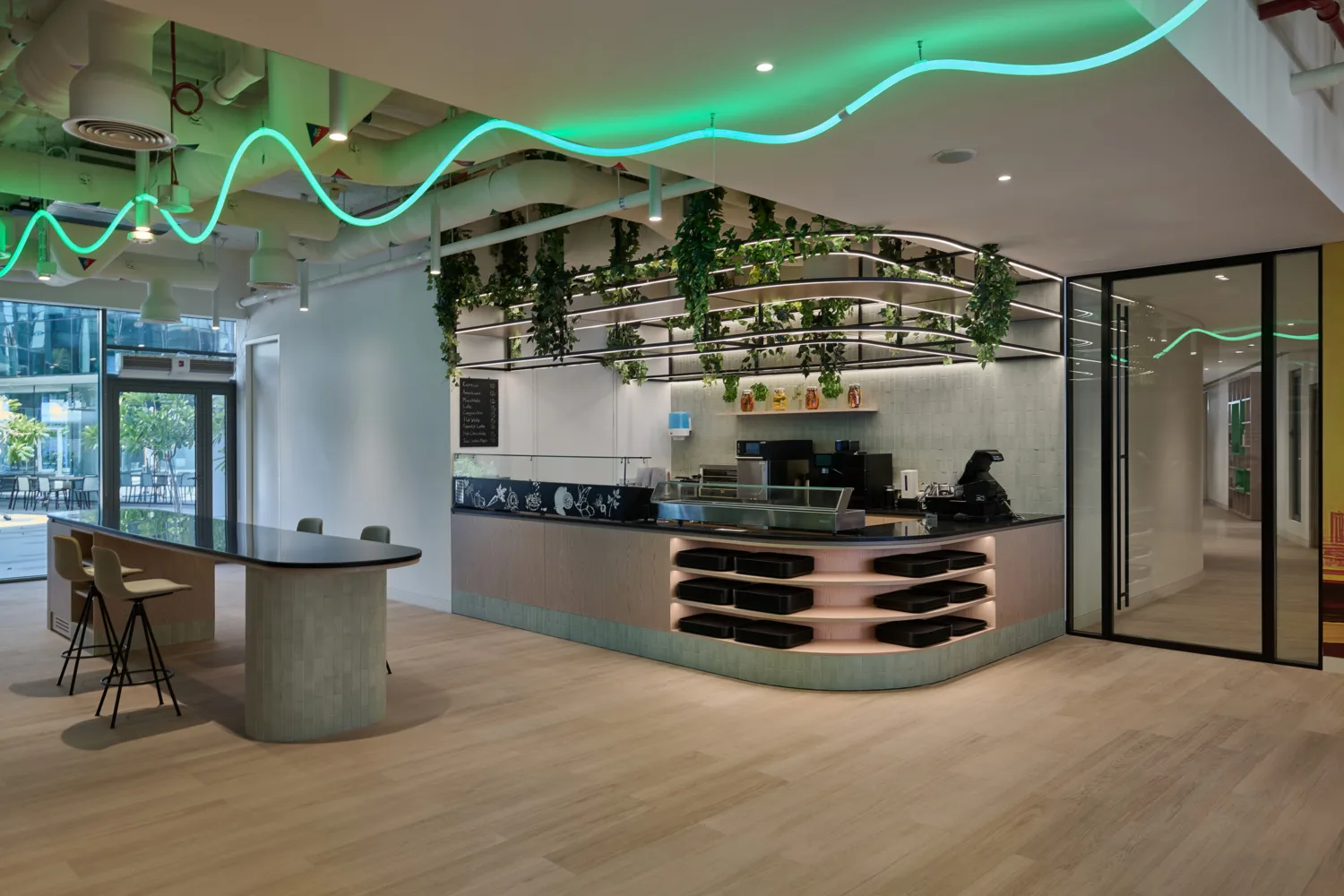 Modern café interior with a counter on the right, hanging plants, shelves with items, and a blackboard menu. A long table with stools is on the left. Ceiling features exposed pipes and green neon lights.