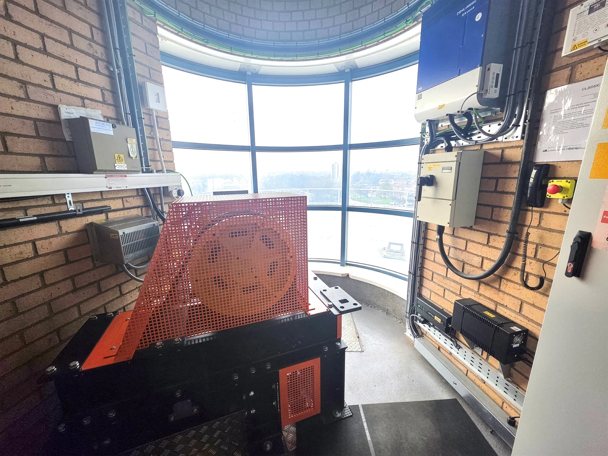 A small machinery room with exposed brick walls contains a large orange‑cased mechanical unit on the floor, surrounded by electrical panels, cables, and conduits. A curved window provides a bright exterior view.