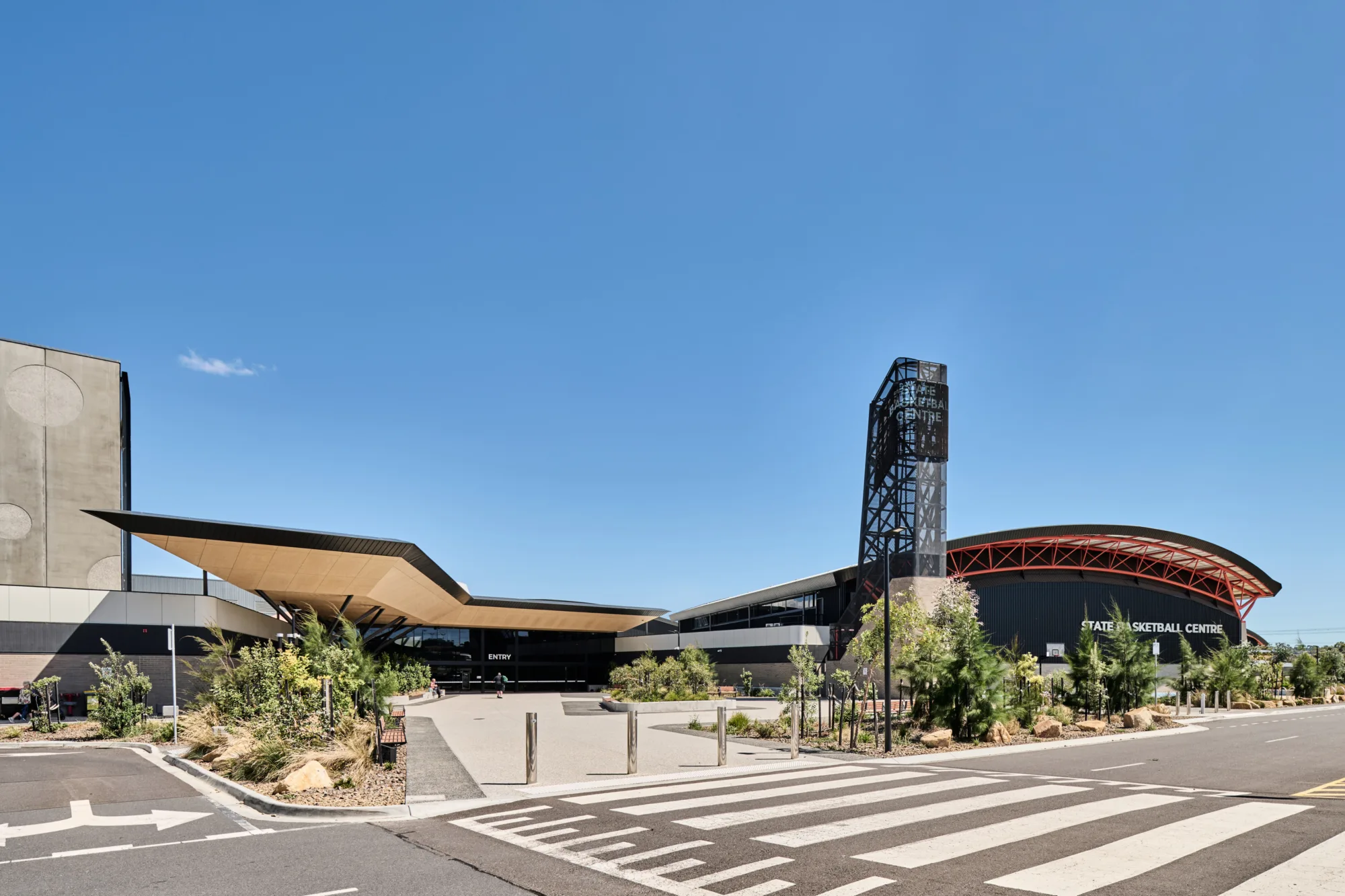 Wide exterior view of a modern sports complex with angular roof lines and landscaped gardens. A tower displays “STATE BASKETBALL CENTRE.” An entrance labelled “ENTRY” sits beneath a canopy, with a crosswalk and road in the foreground.