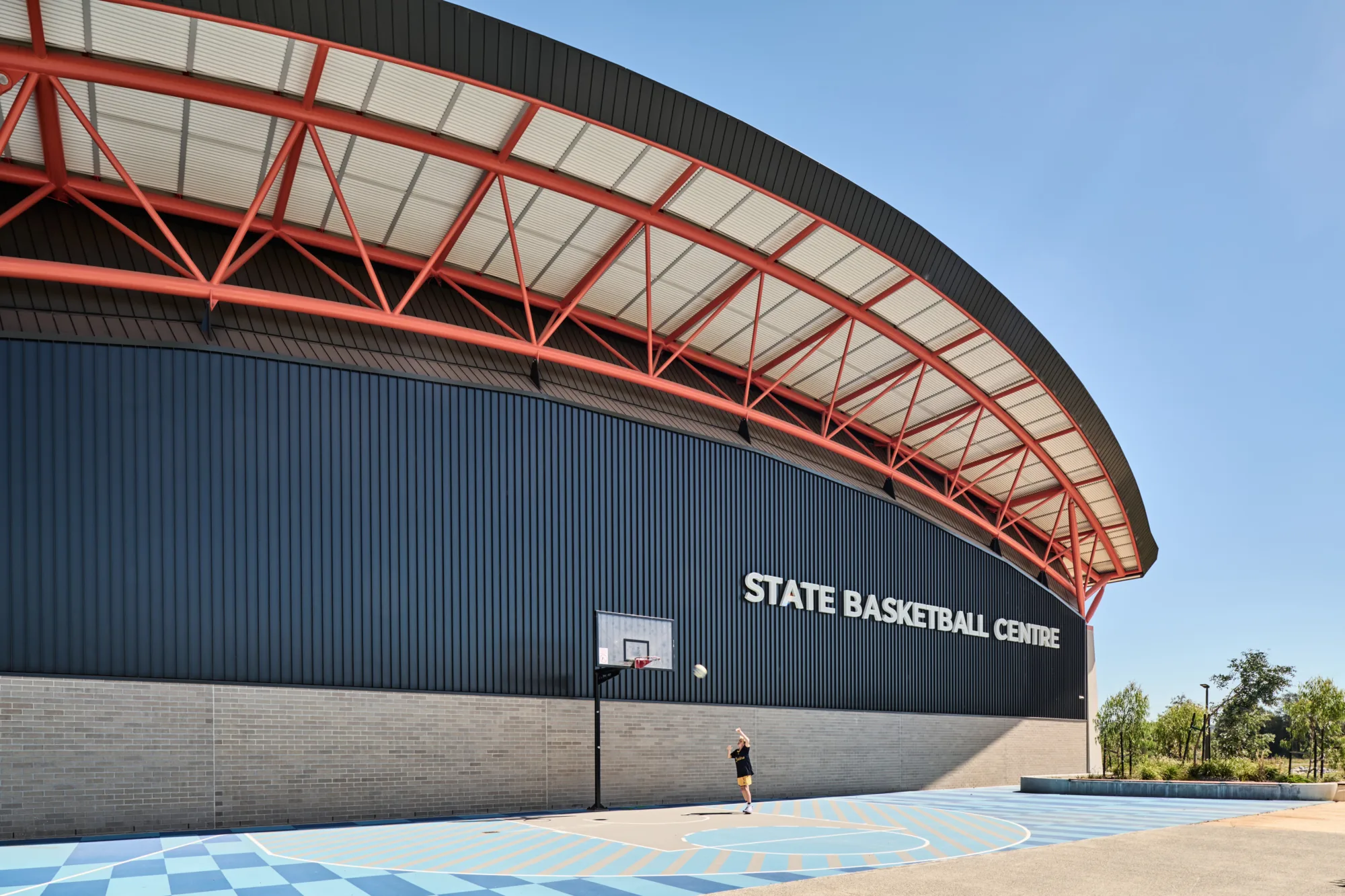 Exterior of a large sports building with a curved roof supported by red steel beams. The facade reads “STATE BASKETBALL CENTRE.” A basketball hoop stands on a blue court where one person is reaching toward a ball mid-air.