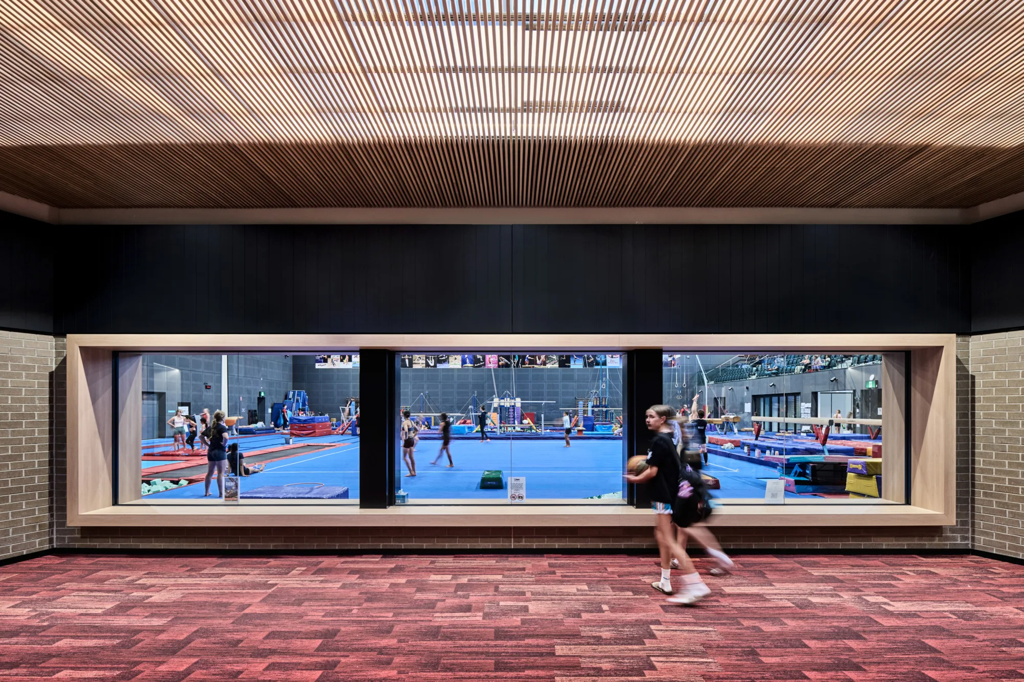 Interior corridor with red patterned carpet and wood slat ceiling. Large windows reveal a gym with blue mats and equipment where several people practice. Two people walk past the glass; small safety notices are posted below.