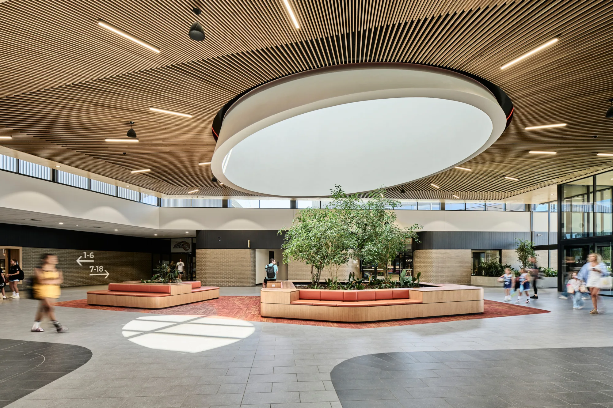 Interior atrium with slatted wood ceiling and large circular skylight fixture above central seating. Curved benches surround planted trees. Several people walk across tiled floor. Wall signage shows arrows with “1–6” and “7–18".
