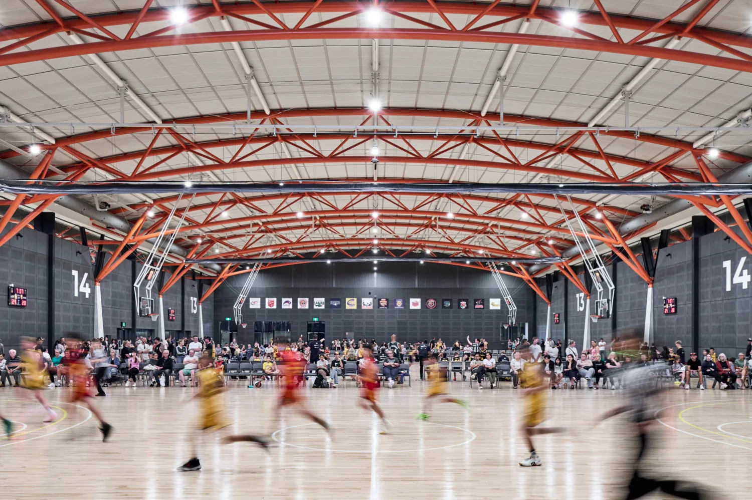 Indoor basketball court with polished wood floor and red steel roof trusses. Blurred players run across the foreground, while rows of seated spectators line the sideline. Large wall numbers “13” and “14” and scoreboards are visible.