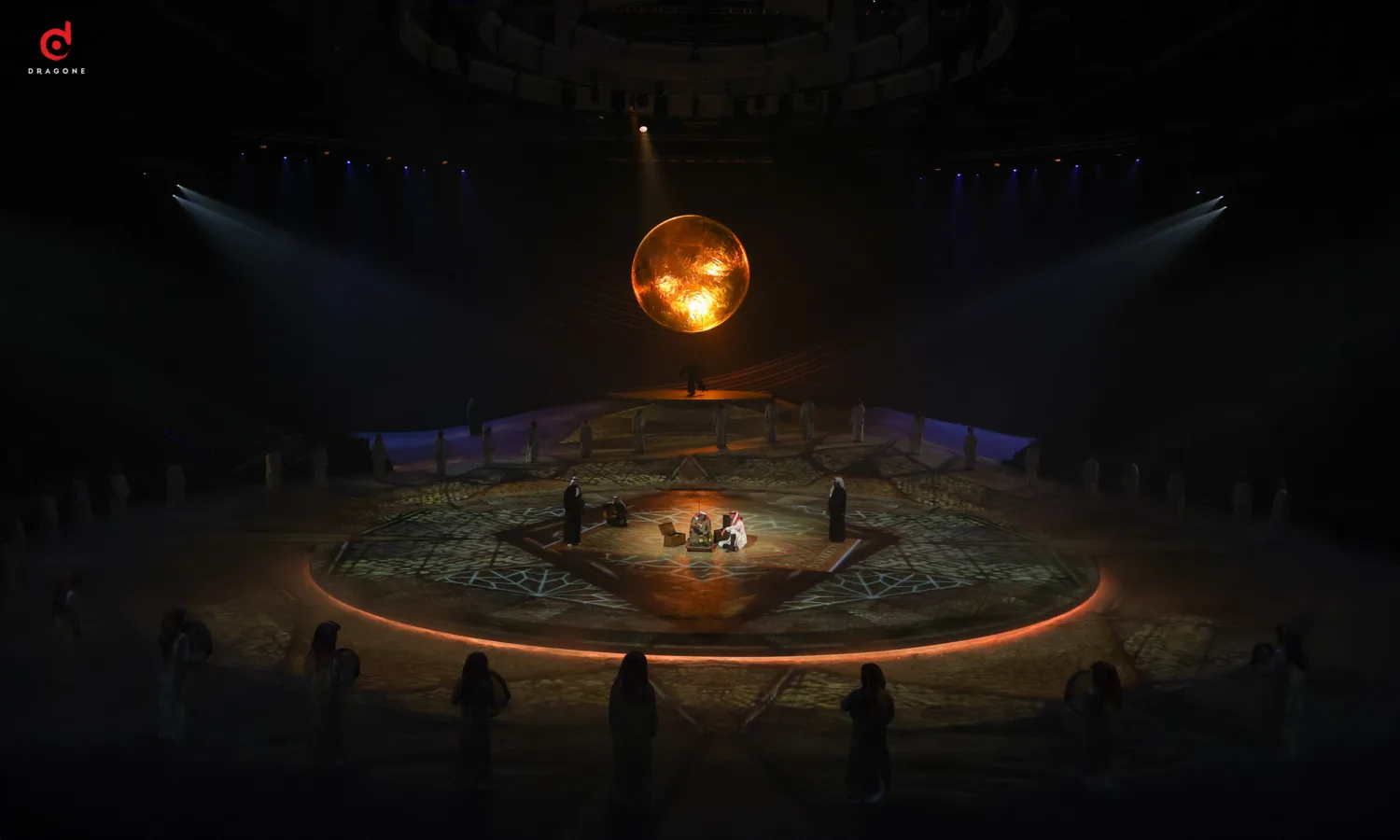 Orange sun like object hanging from centre of stage with performers around