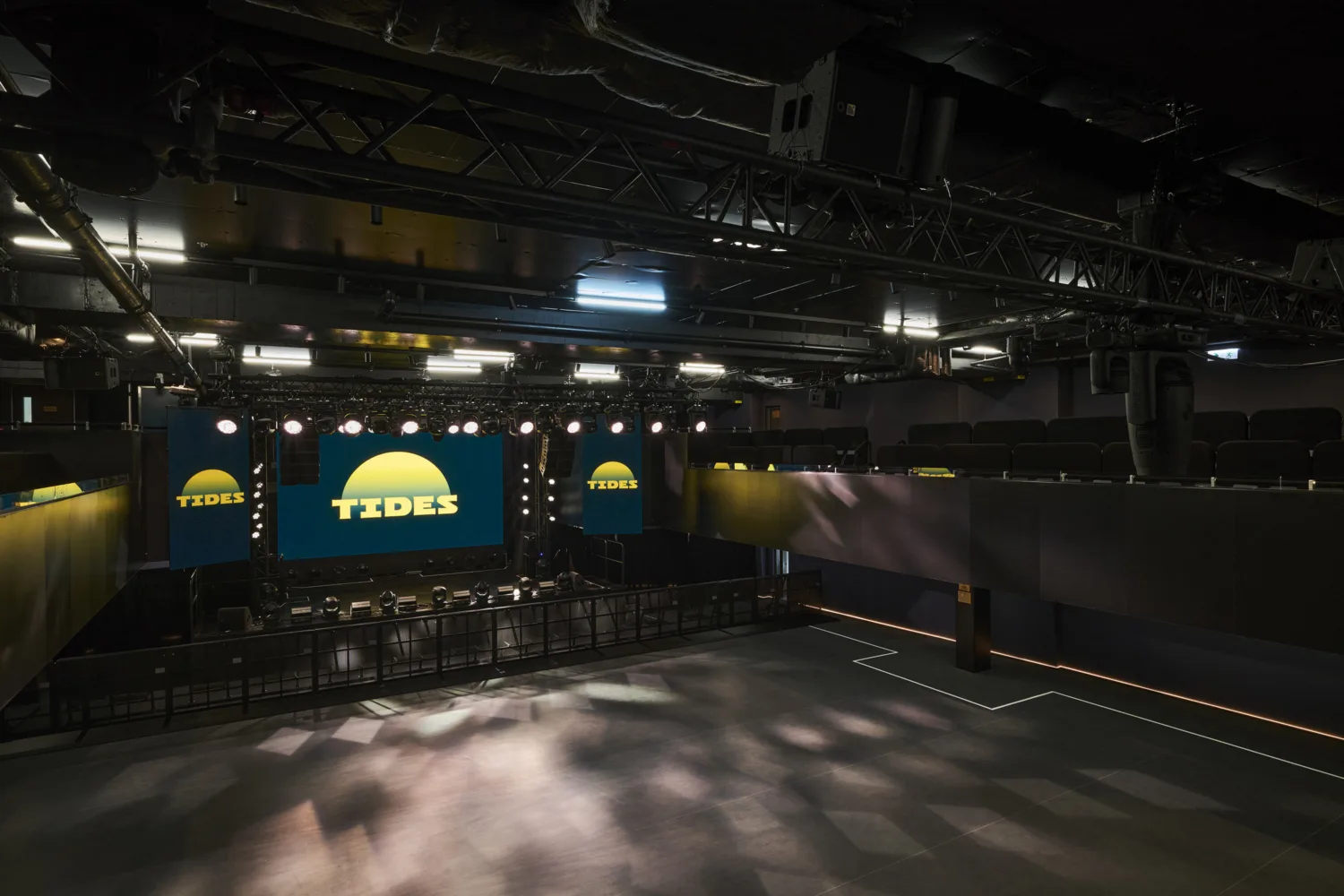A large indoor venue features a wide stage with three screens displaying a yellow sun‑shaped graphic and the word “TIDES.” Overhead rigging, lighting equipment, seating areas, and an empty open floor are visible.