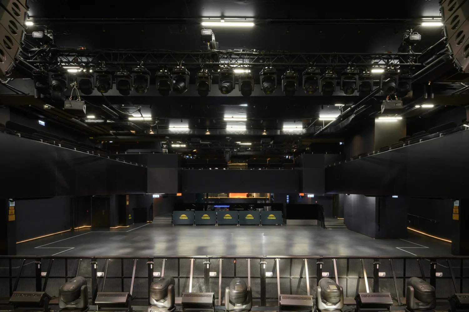 A large indoor venue with an empty open floor faces a bar area at the back displaying three screens showing a yellow sun‑shaped graphic and the word “TIDES.” Overhead rigging, lighting equipment, and tiered seating surround the space.