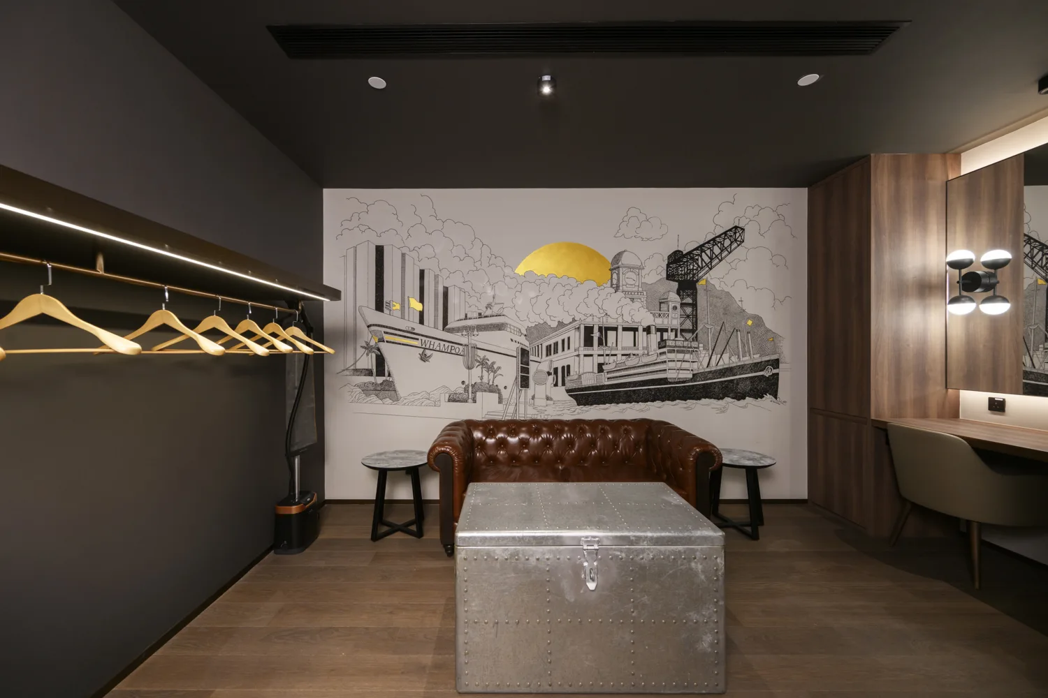 Modern interior room with a brown leather sofa centered beneath a wall mural of ships, buildings, crane, and a yellow sun. Metal trunk table in front, side tables, coat rack with wooden hangers, and lit mirror vanity.