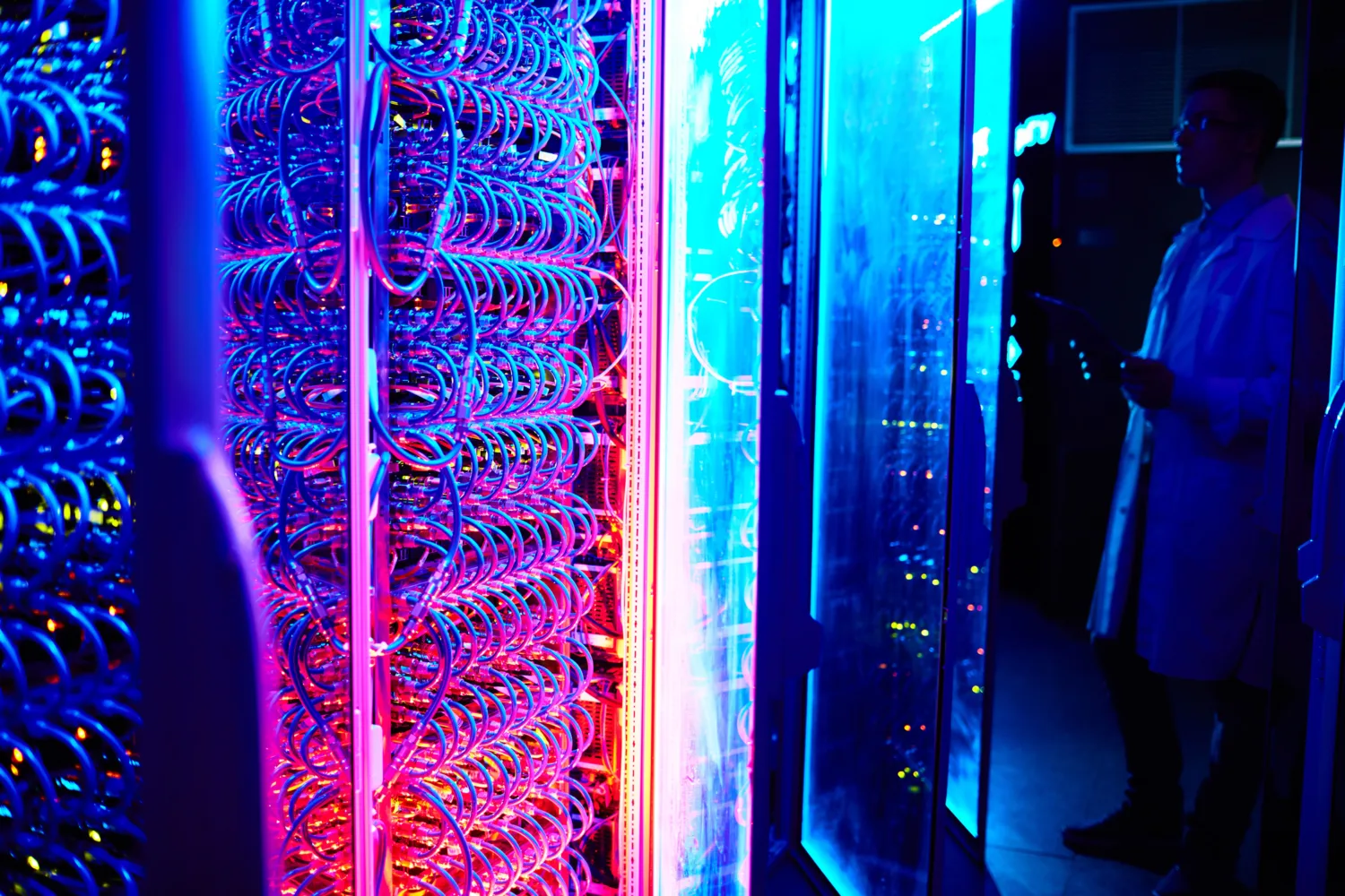 Rows of illuminated liquid cooling loops and equipment inside server racks, with a person standing beside glass-fronted cabinets in a data centre.