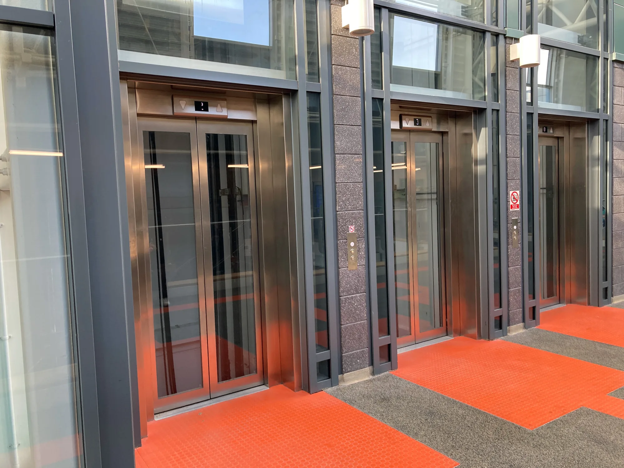 Three glass lift doors with red textured flooring at the entrance