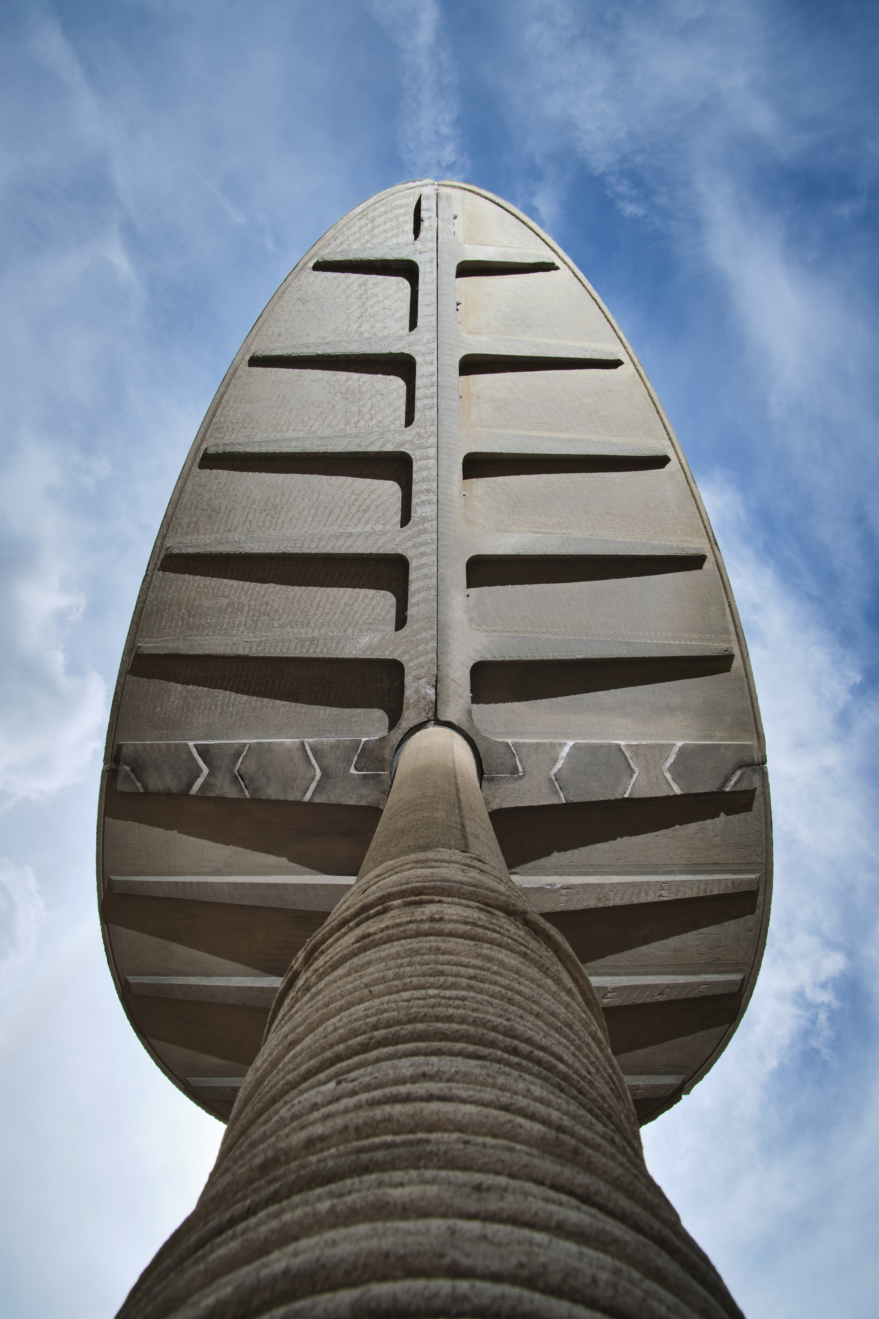 Low-angle view of the Canopy, a large concrete structure with a ribbed stem and segmented, oval-shaped top, set against a blue sky with scattered clouds.