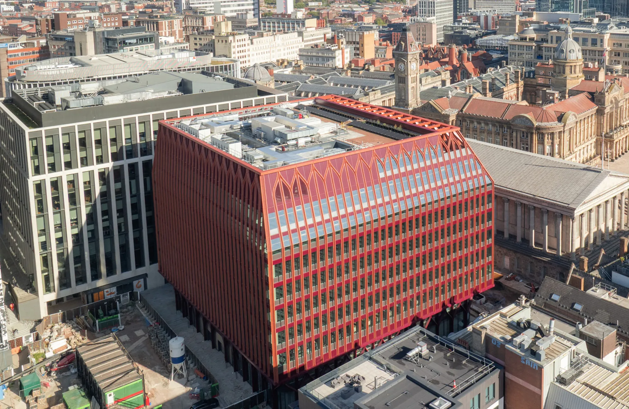 Aerial view of a city block featuring a modern red building with a slanted roof and vertical fins, surrounded by older historic structures and other office buildings. Rooftop equipment is visible, and streets with construction areas are in the foreground.